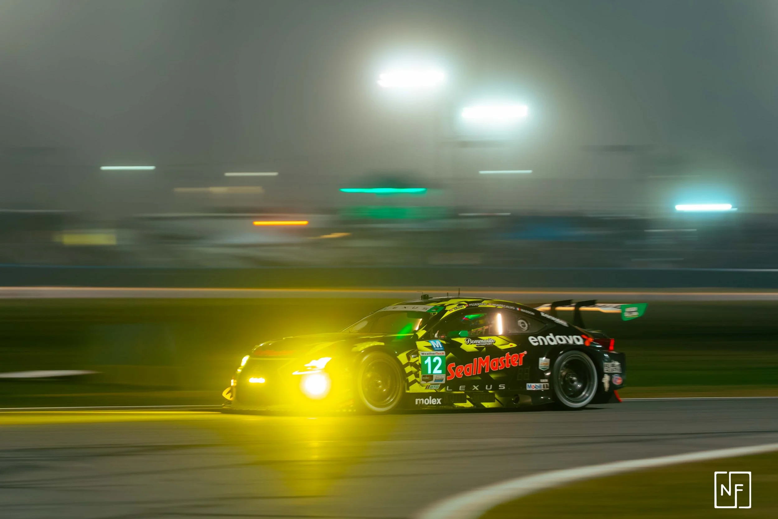 Race car speeding on track at night with motion blur, illuminated headlights, and sponsorship logos.