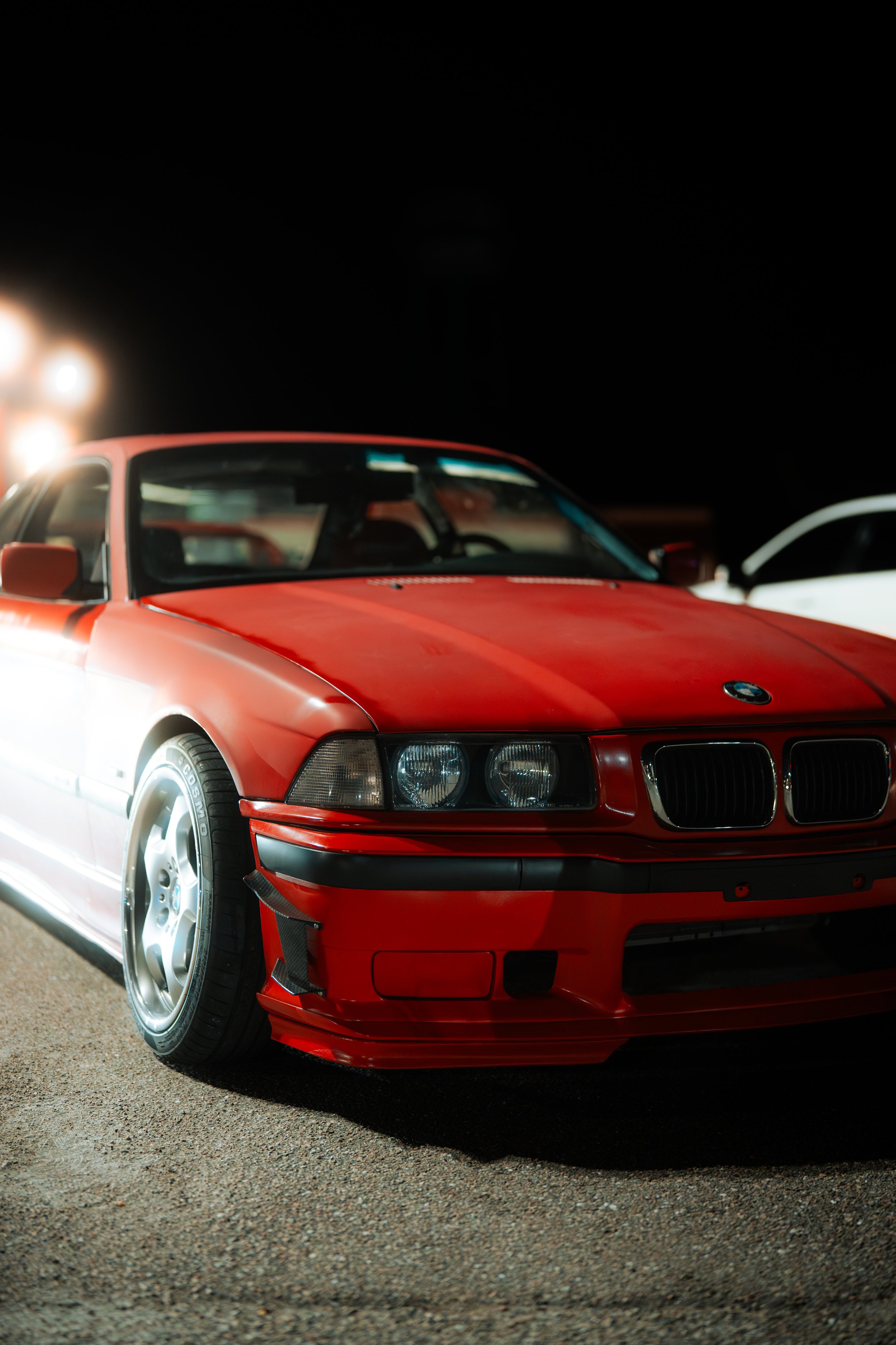 Red BMW E30 parked on a lot at night with bright lights in the background.