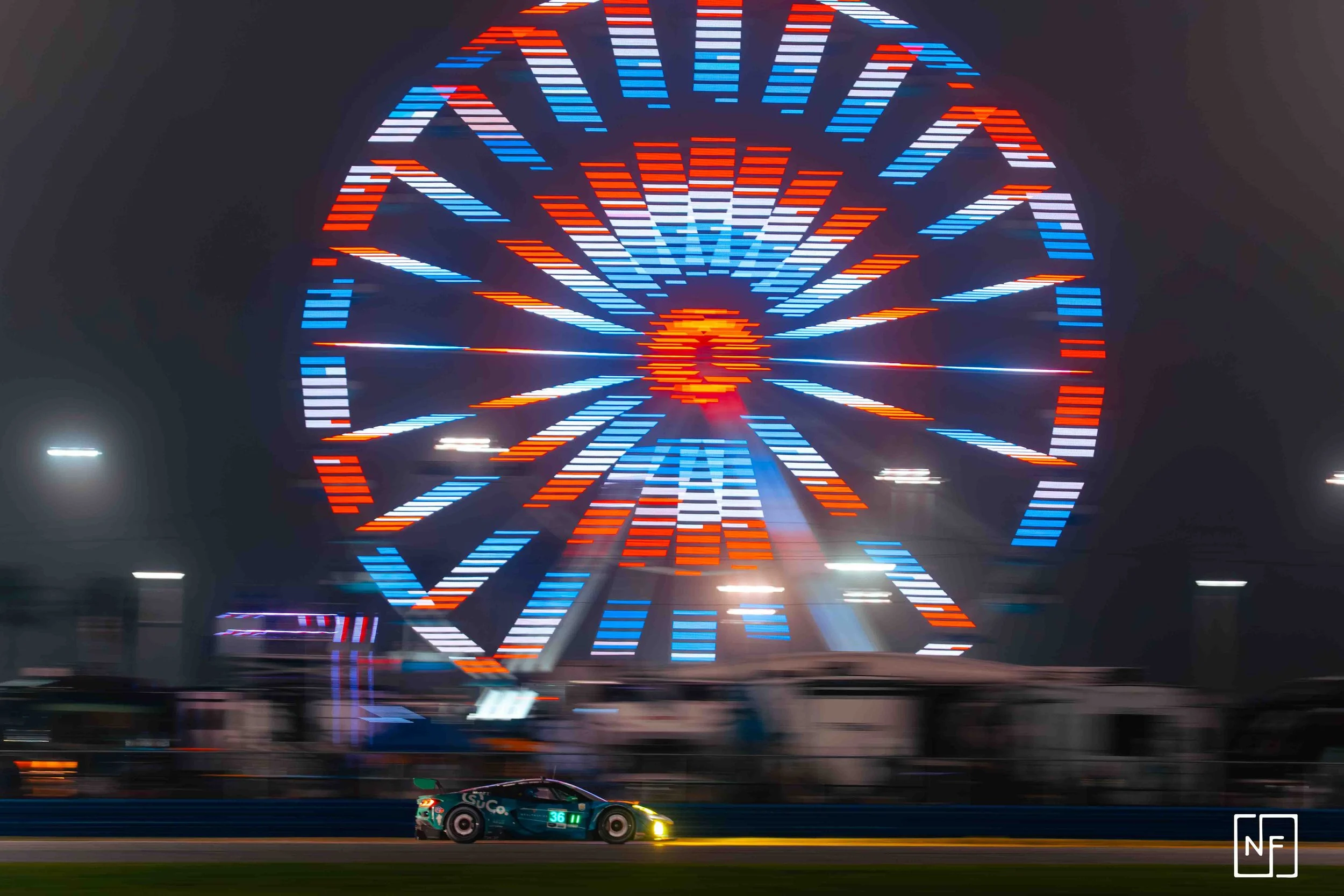 A race car on a track with a large, illuminated Ferris wheel in the background at night with colorful lights.