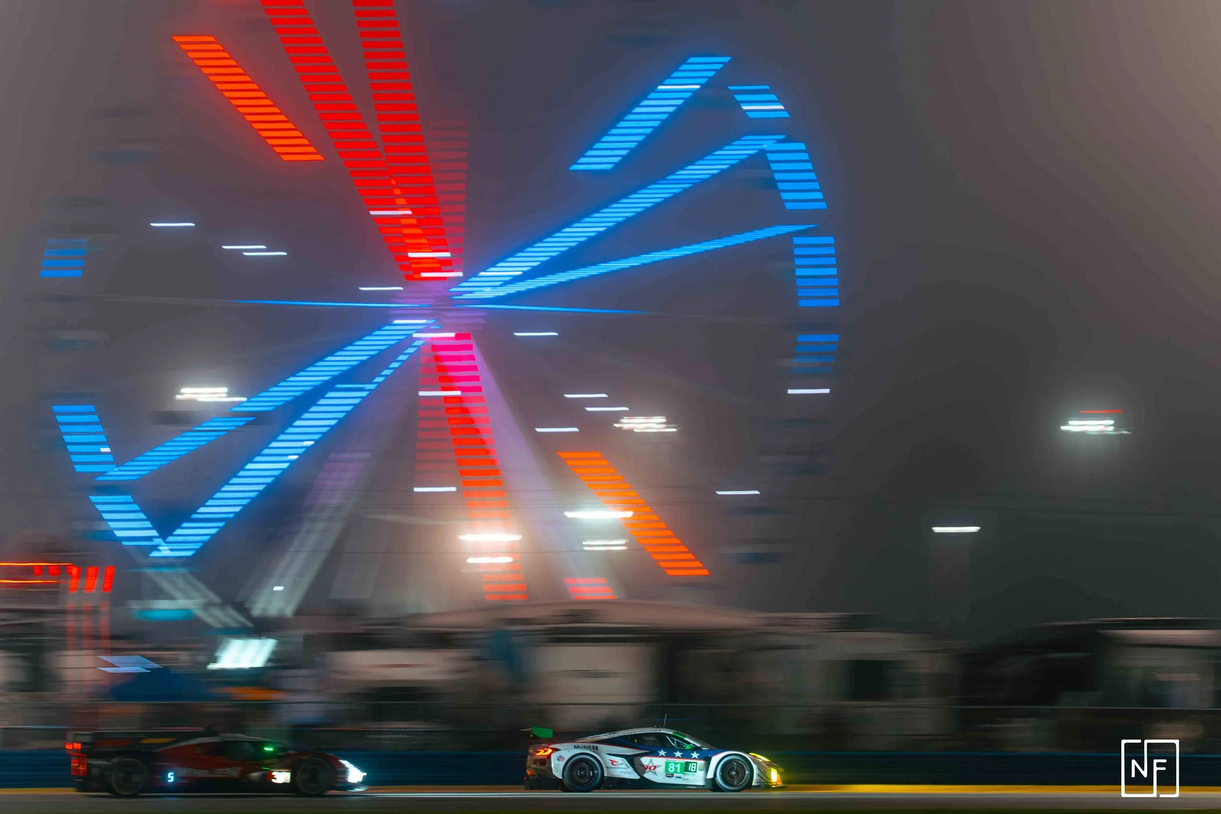 Nighttime scene of a race track with a red and blue neon-lit Ferris wheel in the background. Two race cars, one black and one white, are on the track with motion blur indicating speed.