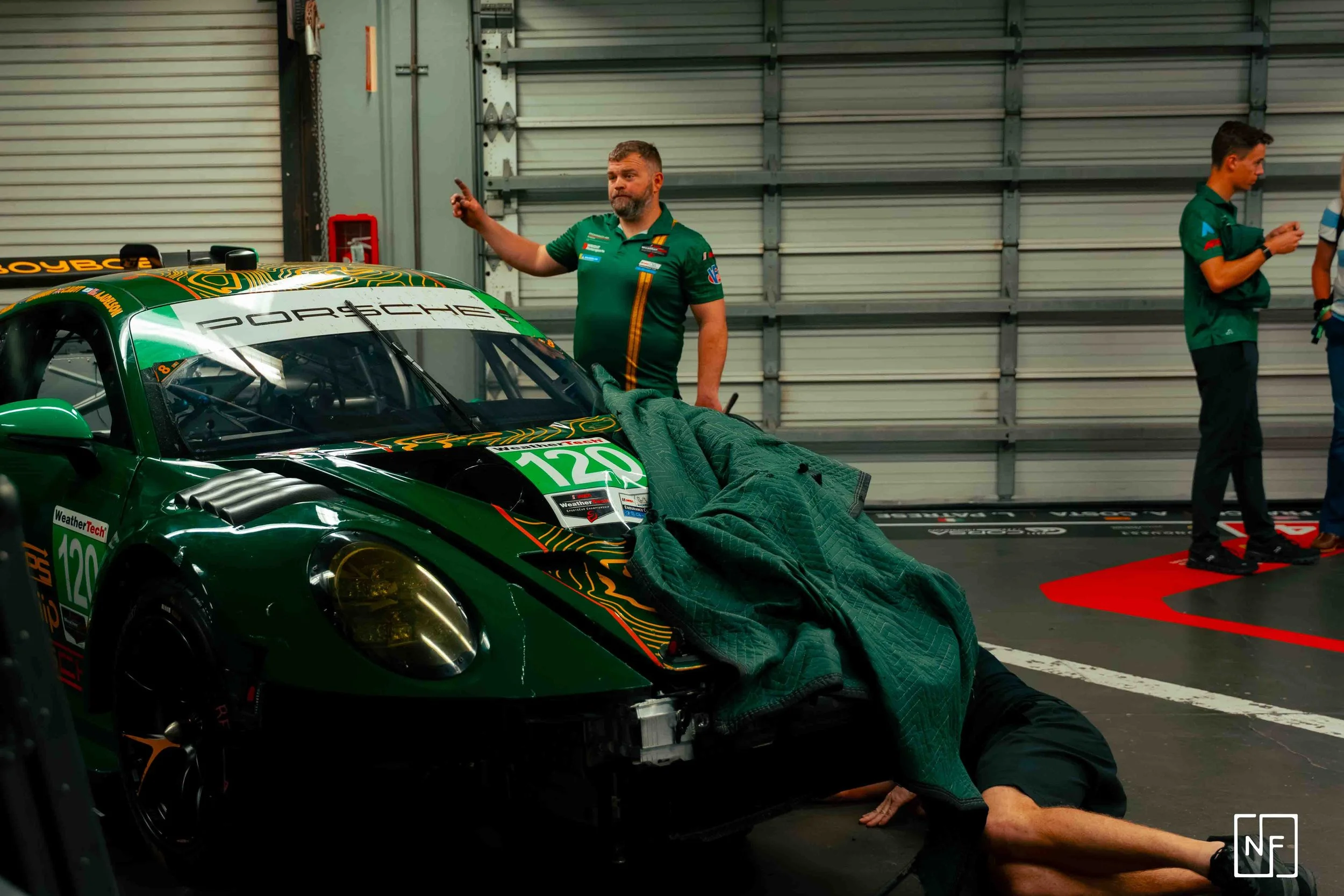 A pit crew member inspecting a green Porsche race car in a garage, with a person working underneath the car covered by a green blanket, while two others stand nearby, one pointing and the other on a phone.