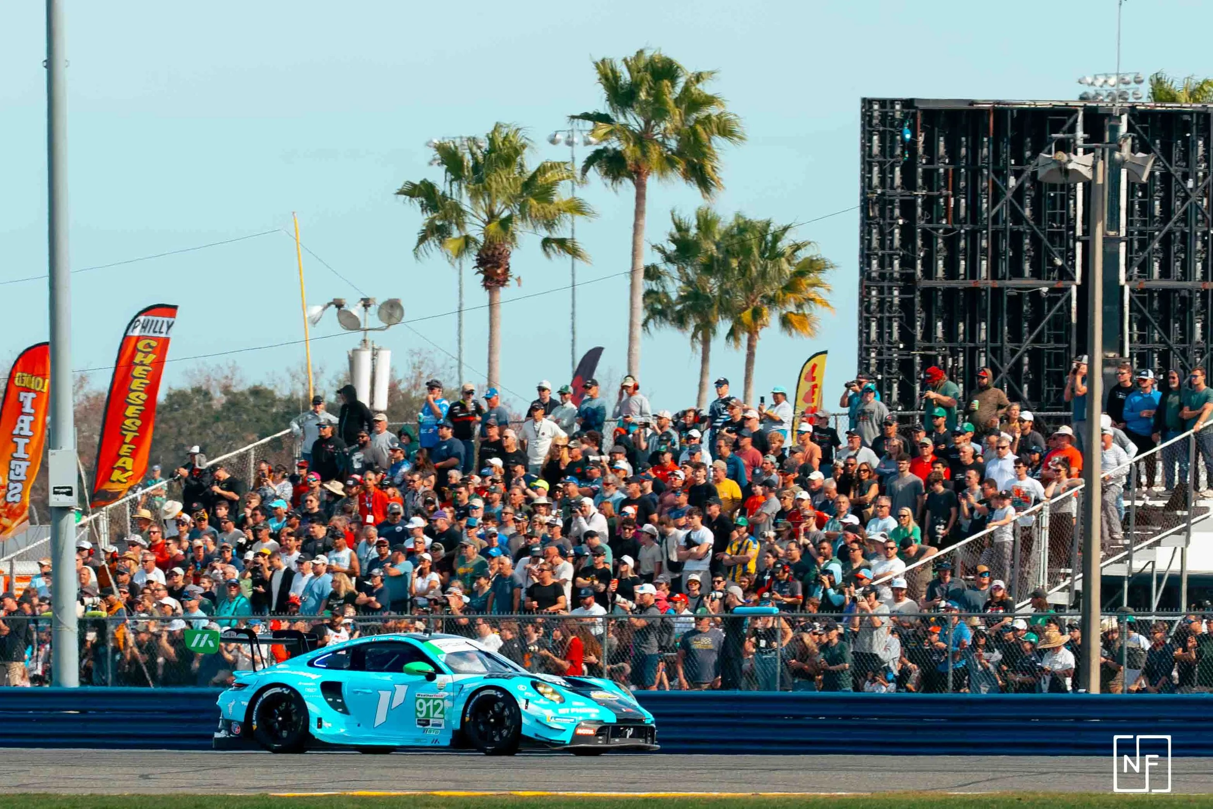 A blue race car on a track with a large crowd of spectators in the background, palm trees, and flags at a racing event.