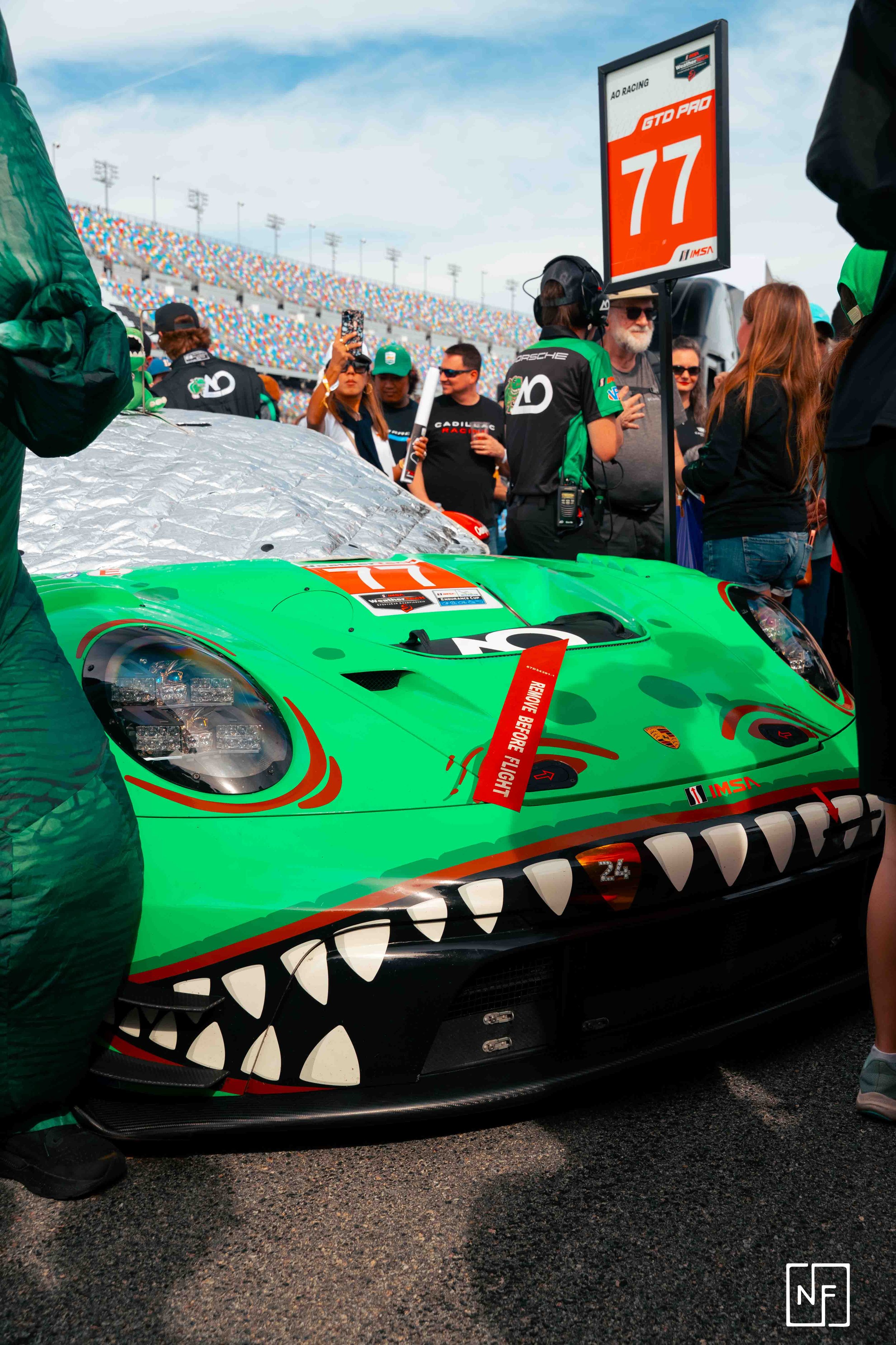 A race car with a green and black shark mouth design on the front, surrounded by spectators and team personnel at a racing event with a grandstand background.