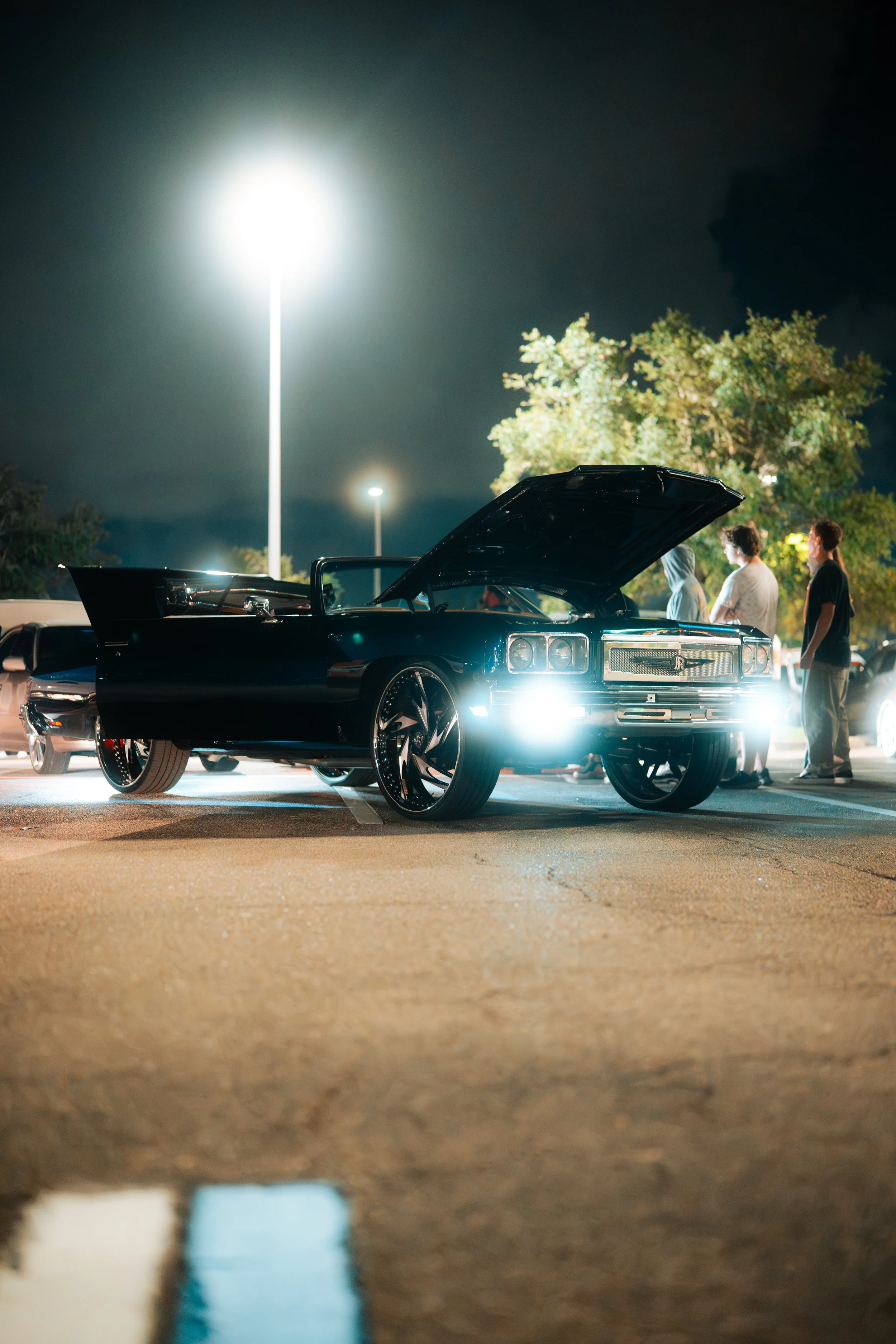 A vintage black car with its hood open parked at night, illuminated by bright headlights, with three people standing nearby and a tree in the background under streetlights.