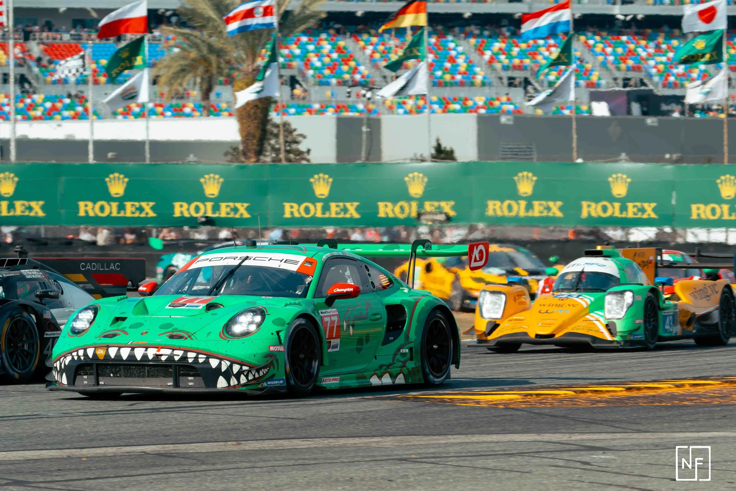 Race car with a front design resembling a crocodile's face, driven by car number 77, racing on a track alongside other race cars with colorful sponsor decals. In the background, flags and banners, including Rolex advertisements, are visible.