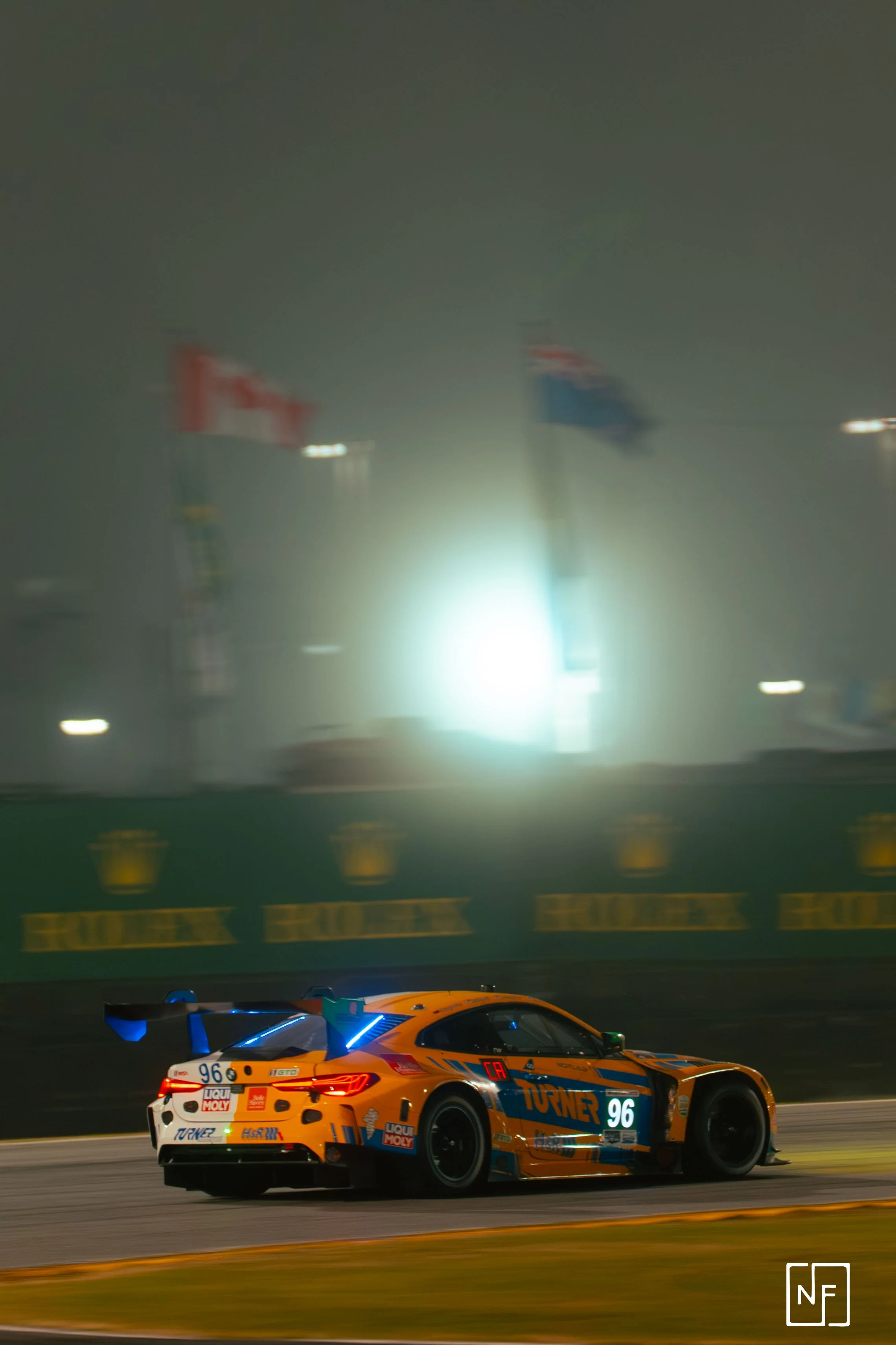 A high-speed race car with an orange, blue, and white livery on a race track at night, with flags and blurred lights in the background.