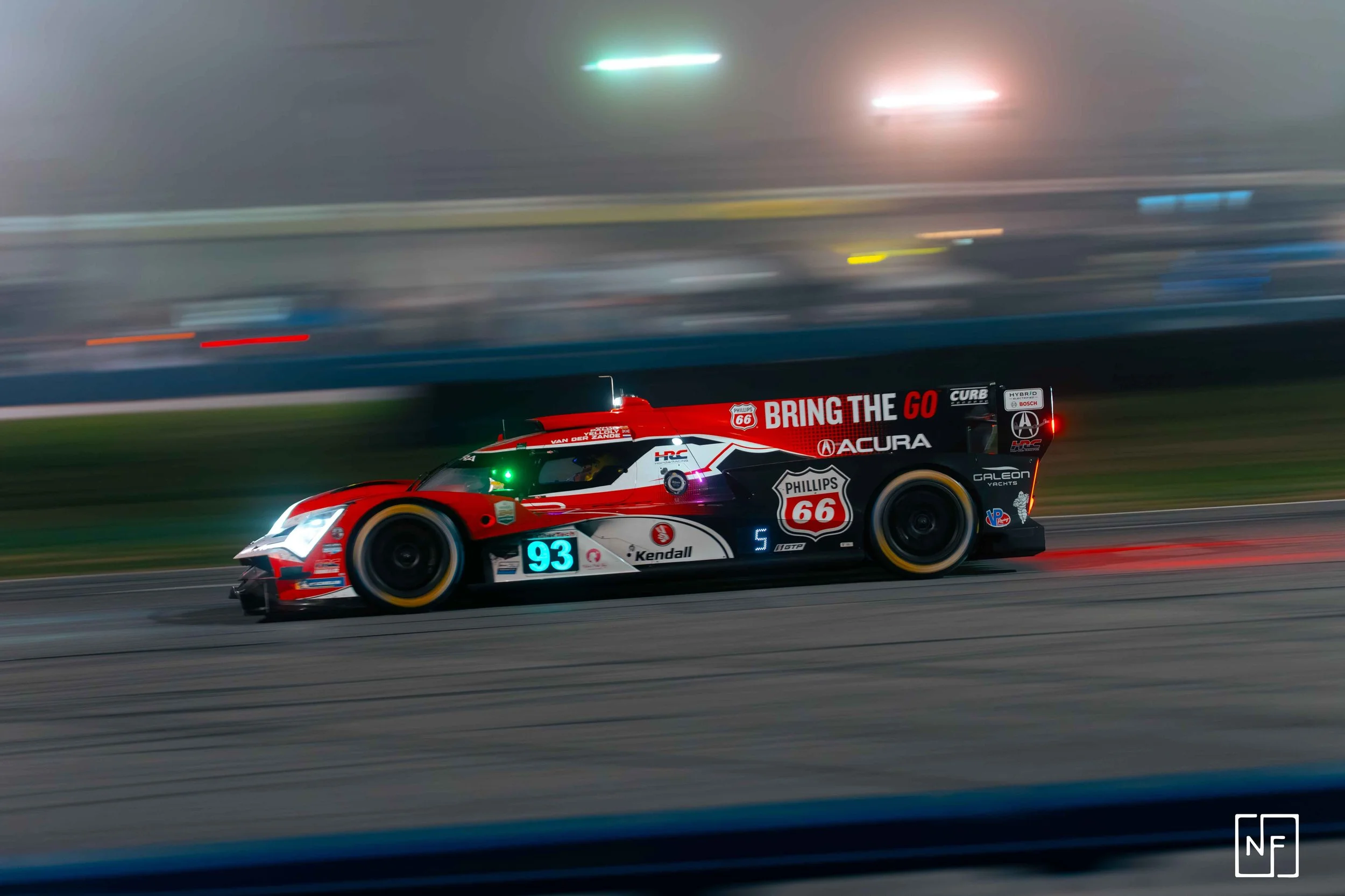 A race car speeding on a track at night, with motion blur in the background. The car is red, white, and black with various sponsor logos, including Phillips 66 and Acura, and the number 66.