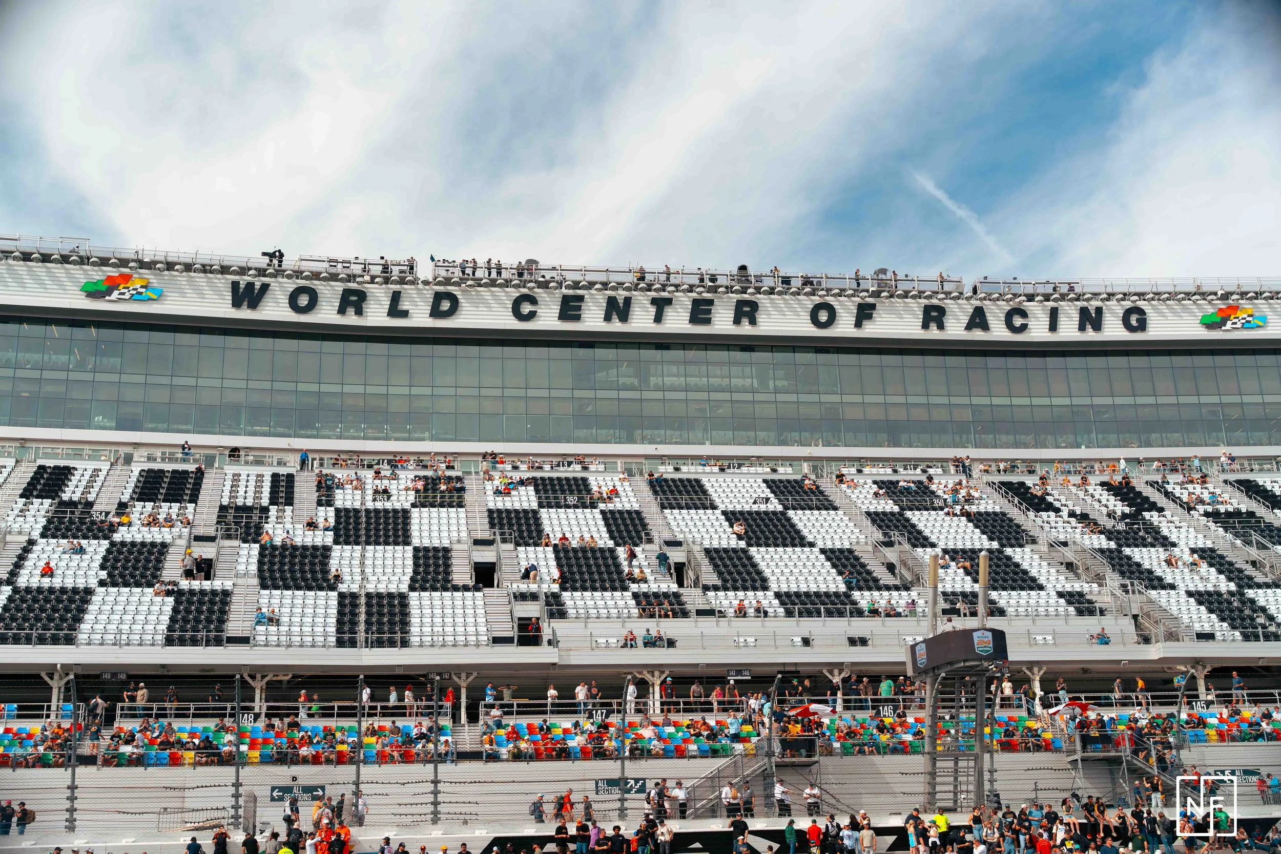 A large racetrack stadium with a sign that reads 'World Center of Racing' on the upper part of the building. Spectator stands filled with people, and the sky is partly cloudy.