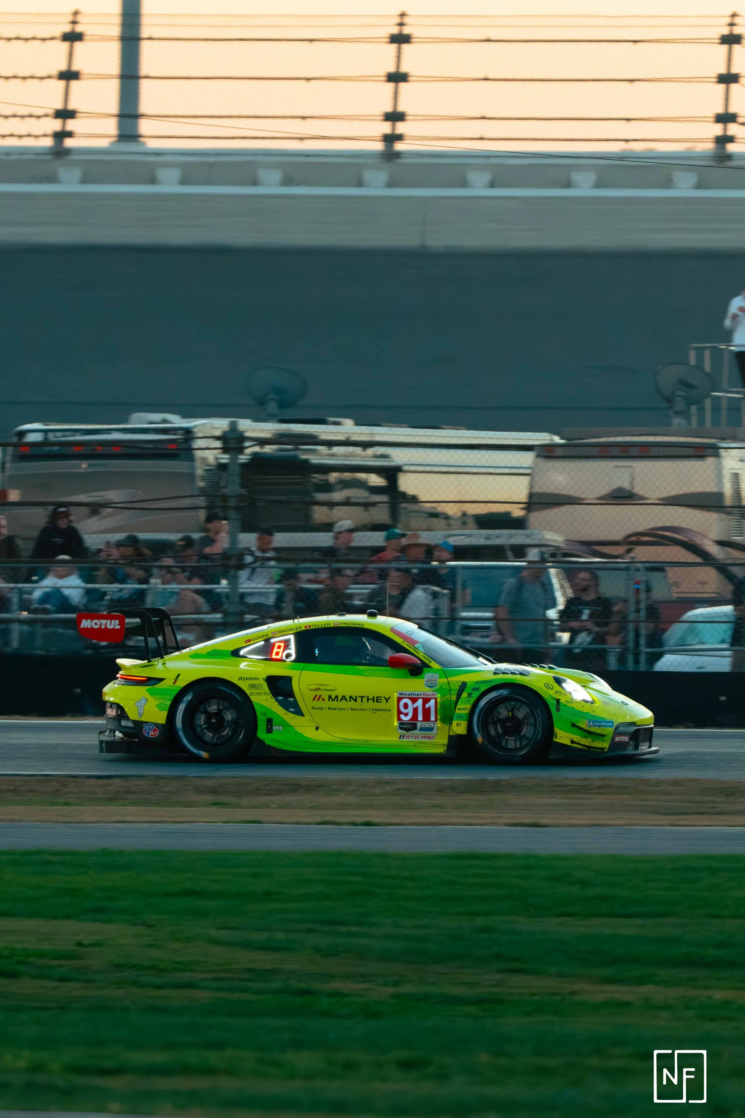 A bright yellow race car with racing decals and the number 911 is driving on a race track, with spectators and trailers in the background during sunset.