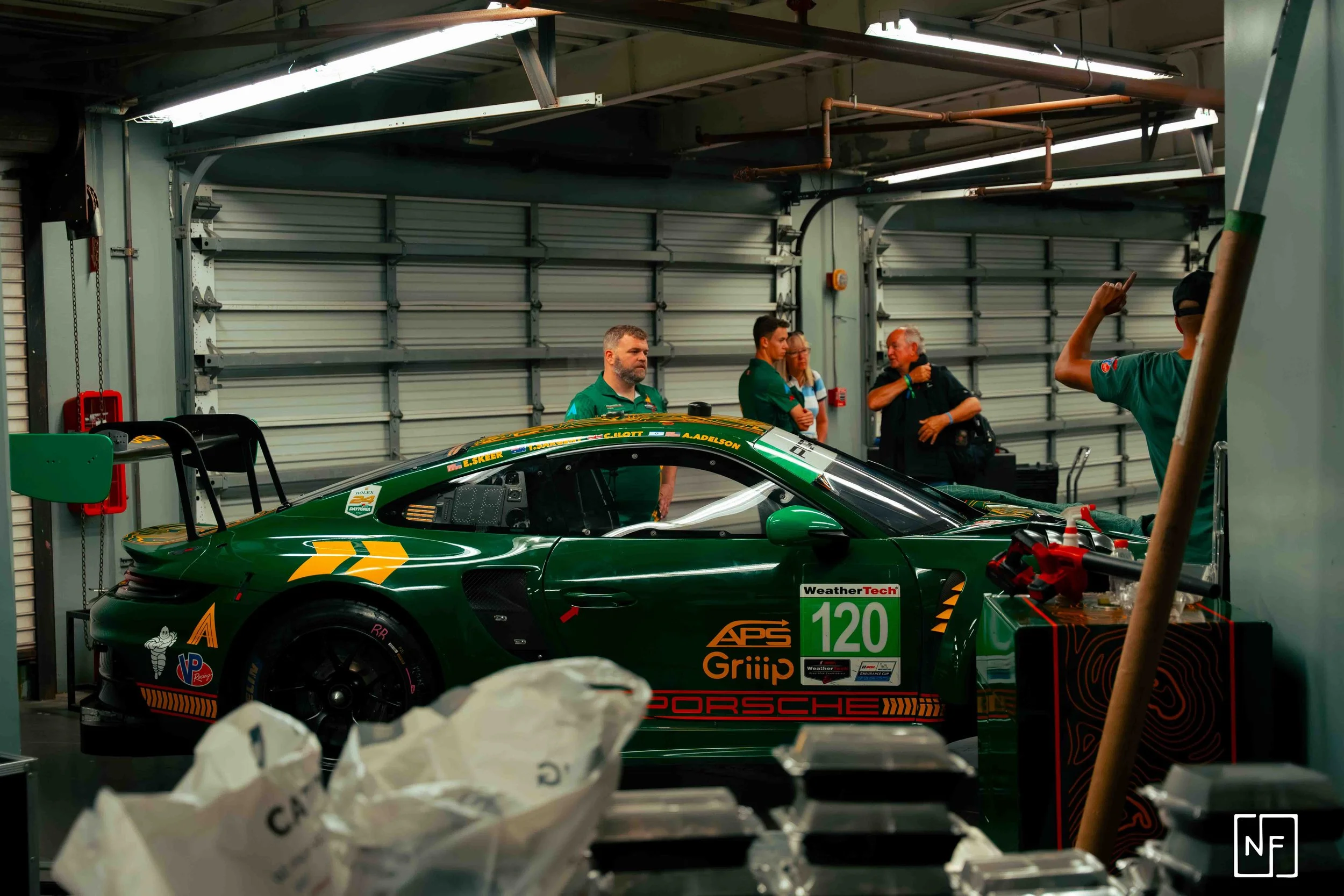 Green race car in a garage with several racing team members standing nearby, engaged in conversation.