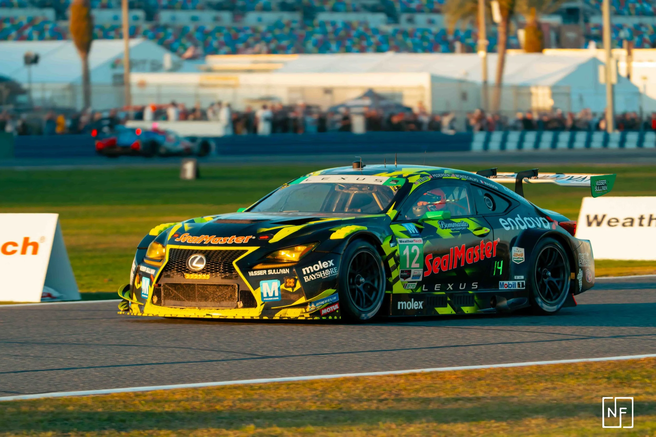 A race car with a yellow and green design on a race track during sunset, sponsored by SealMaster and Lexus.