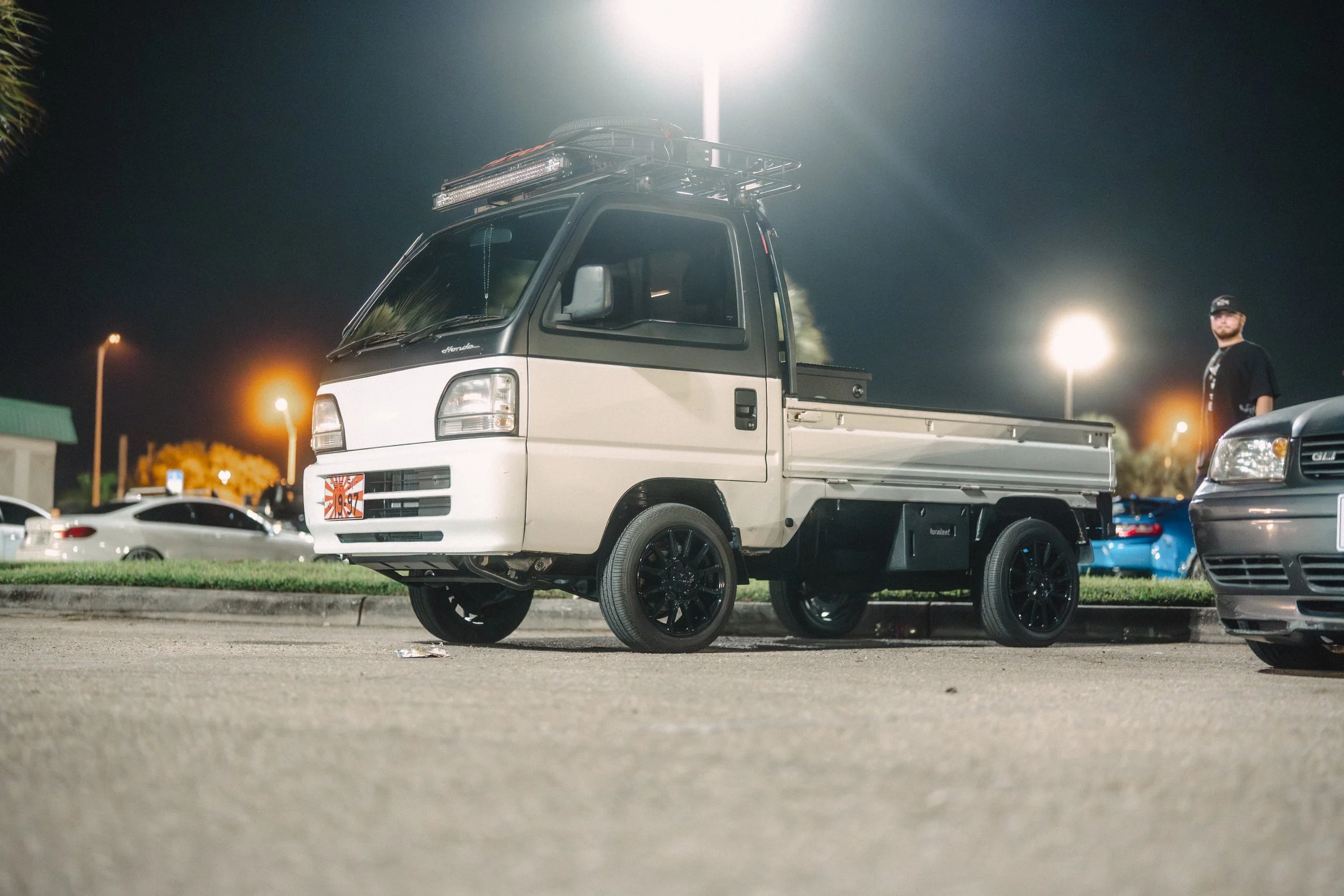 A small white flatbed truck with black wheels parked in a parking lot at night, with a man standing beside a silver car nearby.
