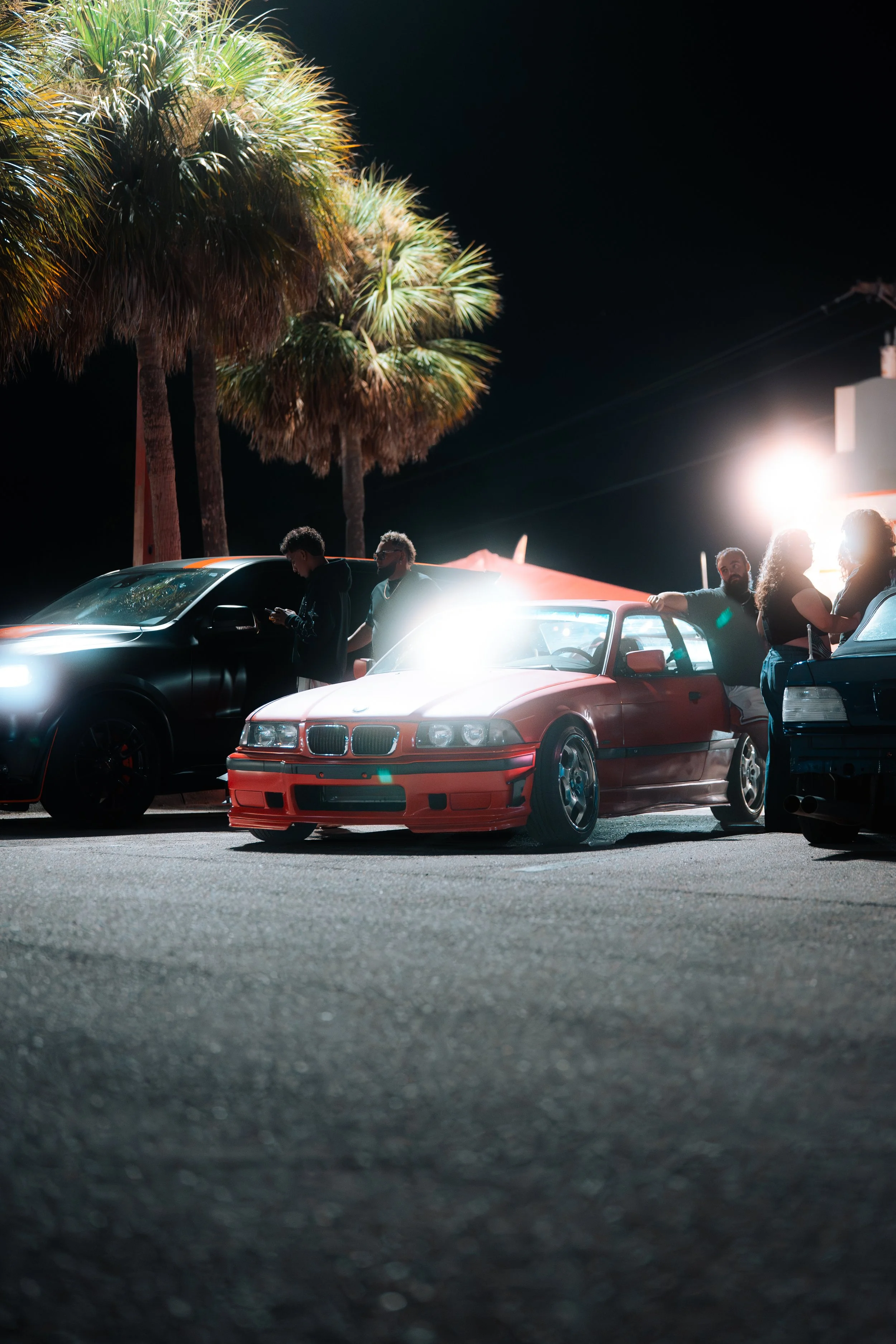 Nighttime scene showing three parked cars, including a red BMW, with four people standing nearby under tall palm trees, illuminated by bright artificial lighting.