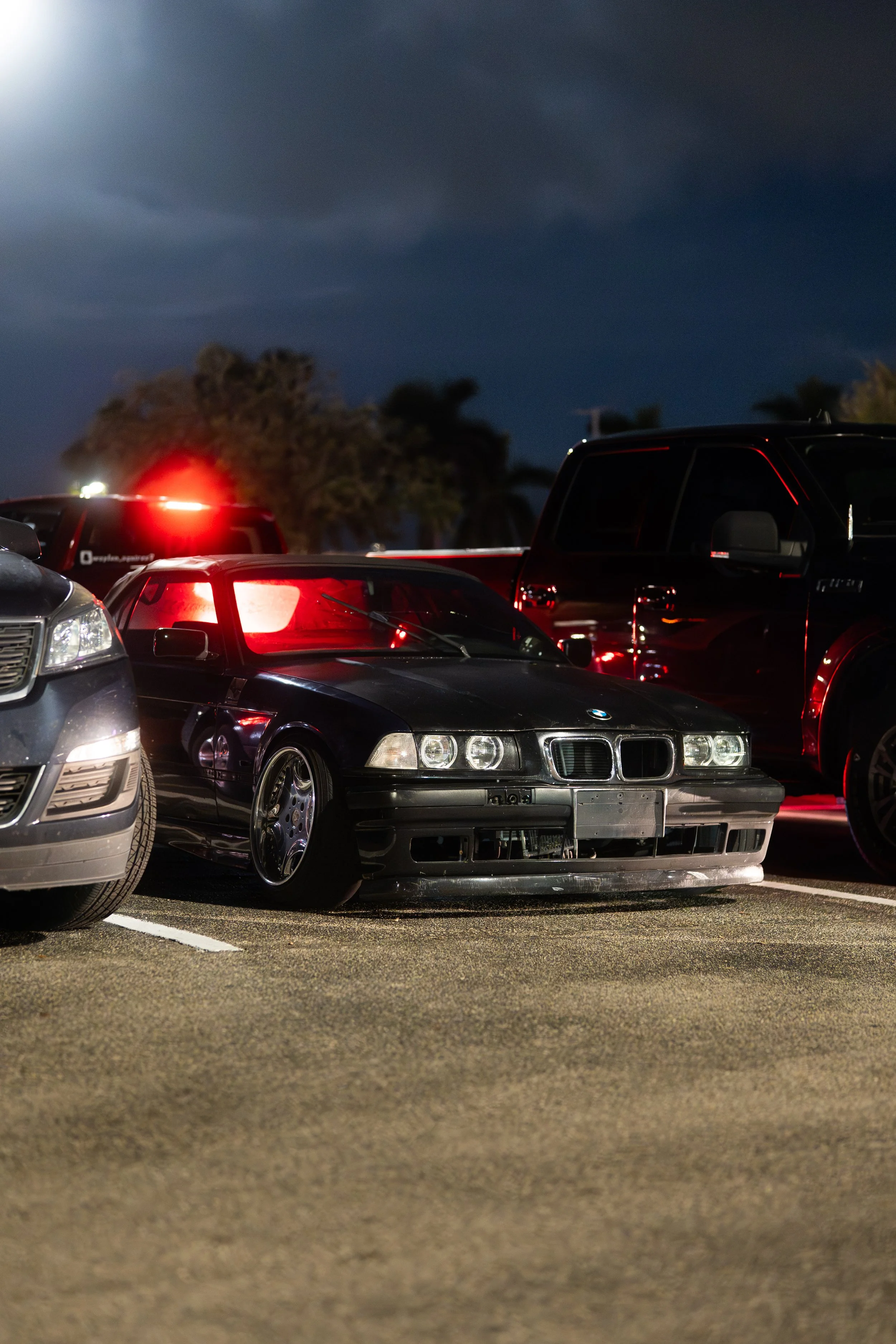 A black vintage BMW Z3 sports car parked in a parking lot at night, surrounded by other vehicles, with trees and a dark sky in the background.