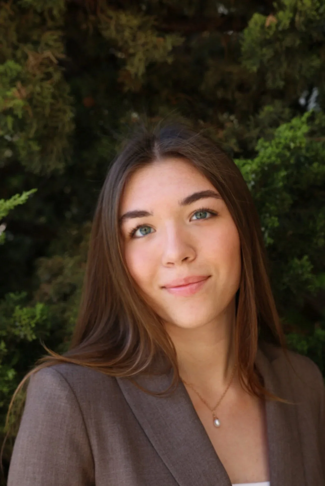 Person with long blonde hair smiling, wearing a dark blazer and a red shirt, standing in front of a building with a large window.