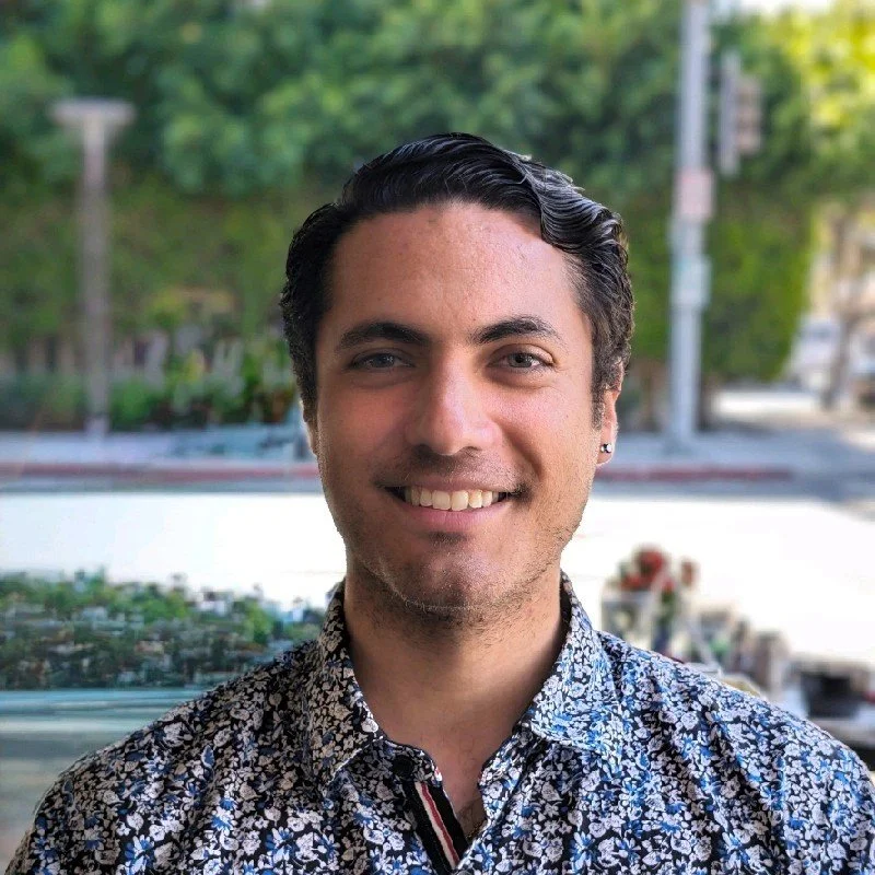 A smiling young man with dark, wavy hair styled back, wearing a floral patterned shirt, standing outdoors with blurred greenery and city street background.