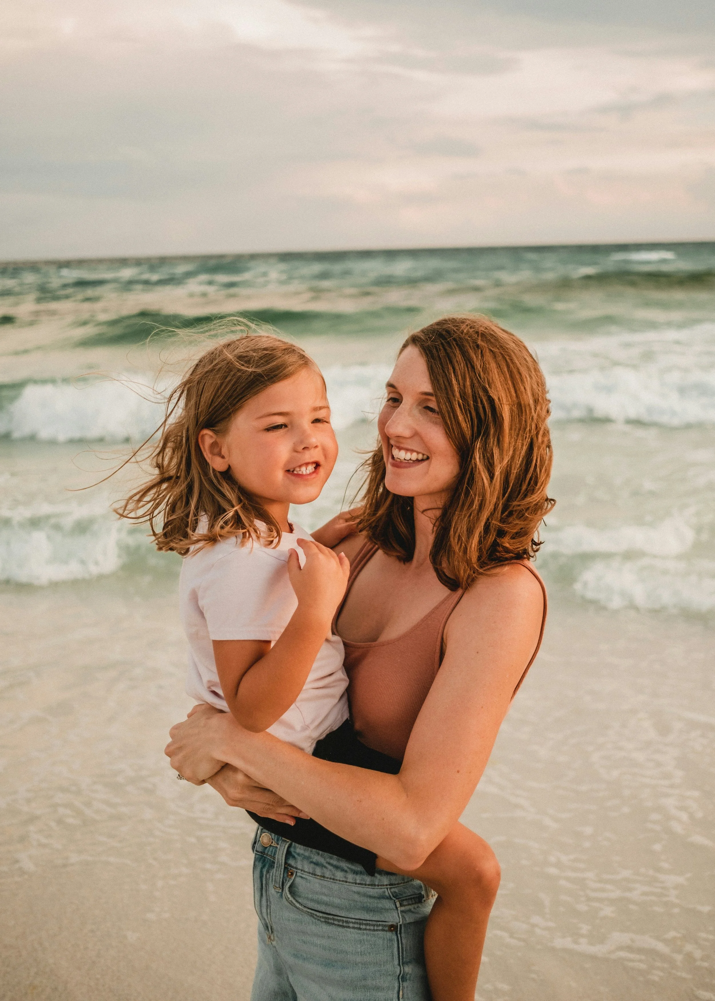 Mother holding child at the beach with ocean waves in the background.