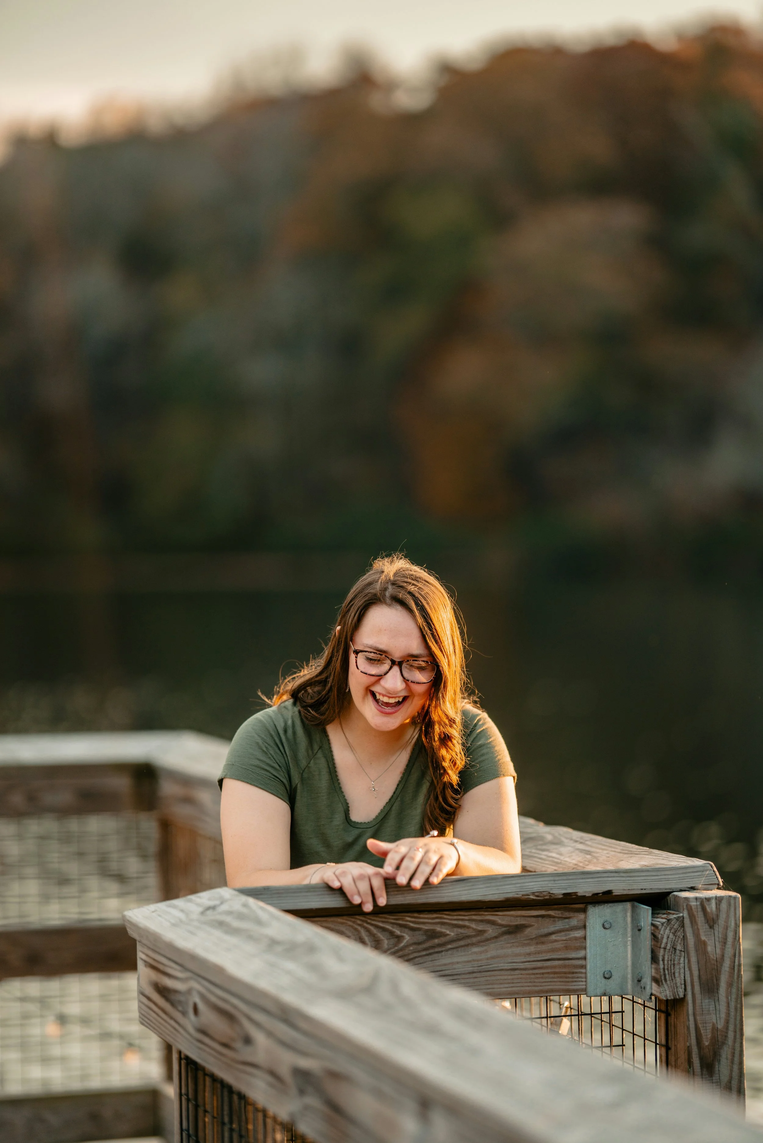 Woman smiling while leaning on a wooden railing by a lake, with blurred trees in the background.