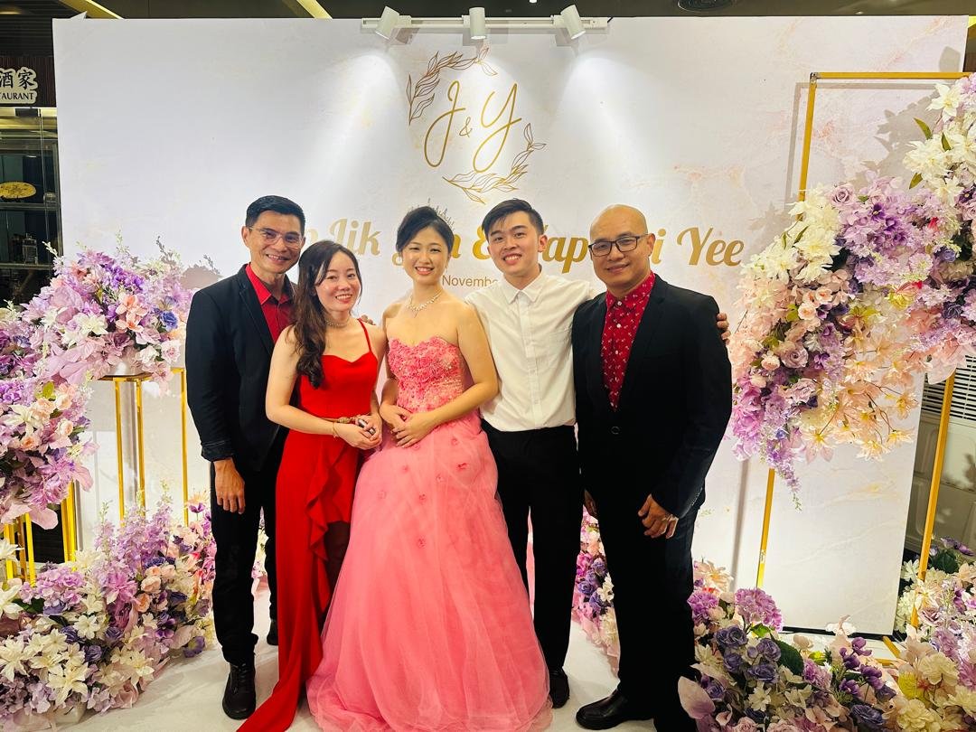 A group of five people posing together at an event with a floral backdrop. Two women are wearing elegant dresses, one in red and the other in pink. Three men are dressed in suits. The backdrop features floral arrangements and decorative initials, sug