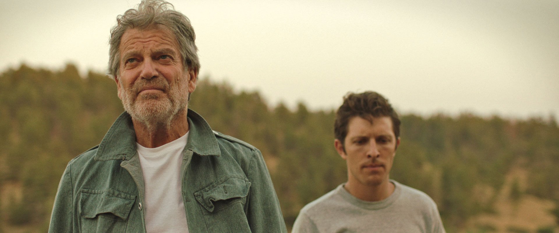 James Storm, wearing a green jacket, with Gabriel Sunday behind him outdoors with a blurred landscape of trees in the background on set for Falcon Song. Directed by Jason Corgan Brown for Corgan Studio. 