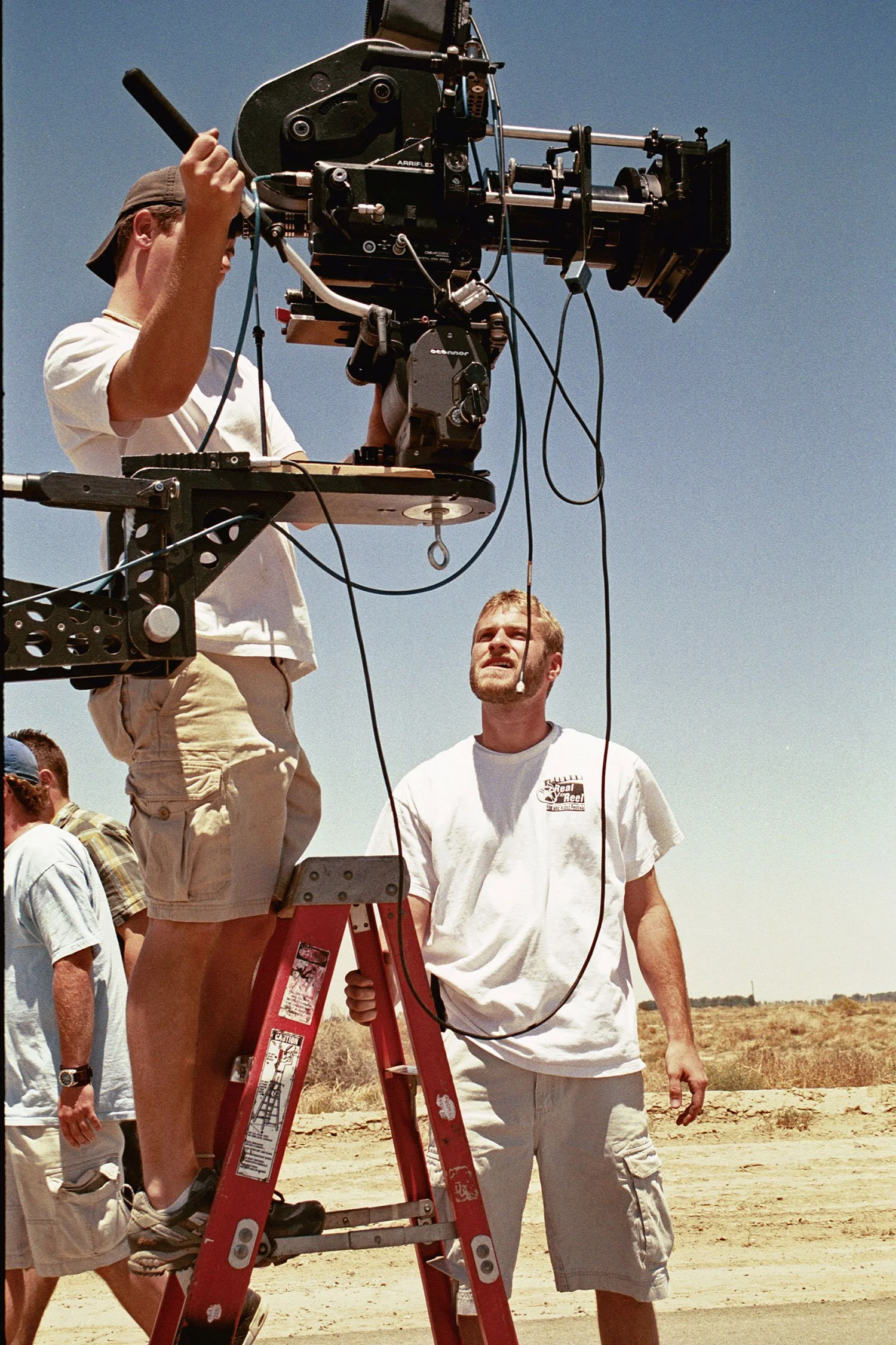 Kenneth Neil Moore setting up a 35mm film camera on a ladder outdoors in a desert-like area during daytime, with clear blue sky and dry landscape in the background. Directed by Ryan Nelson. Produced by Jason Corgan Brown. 