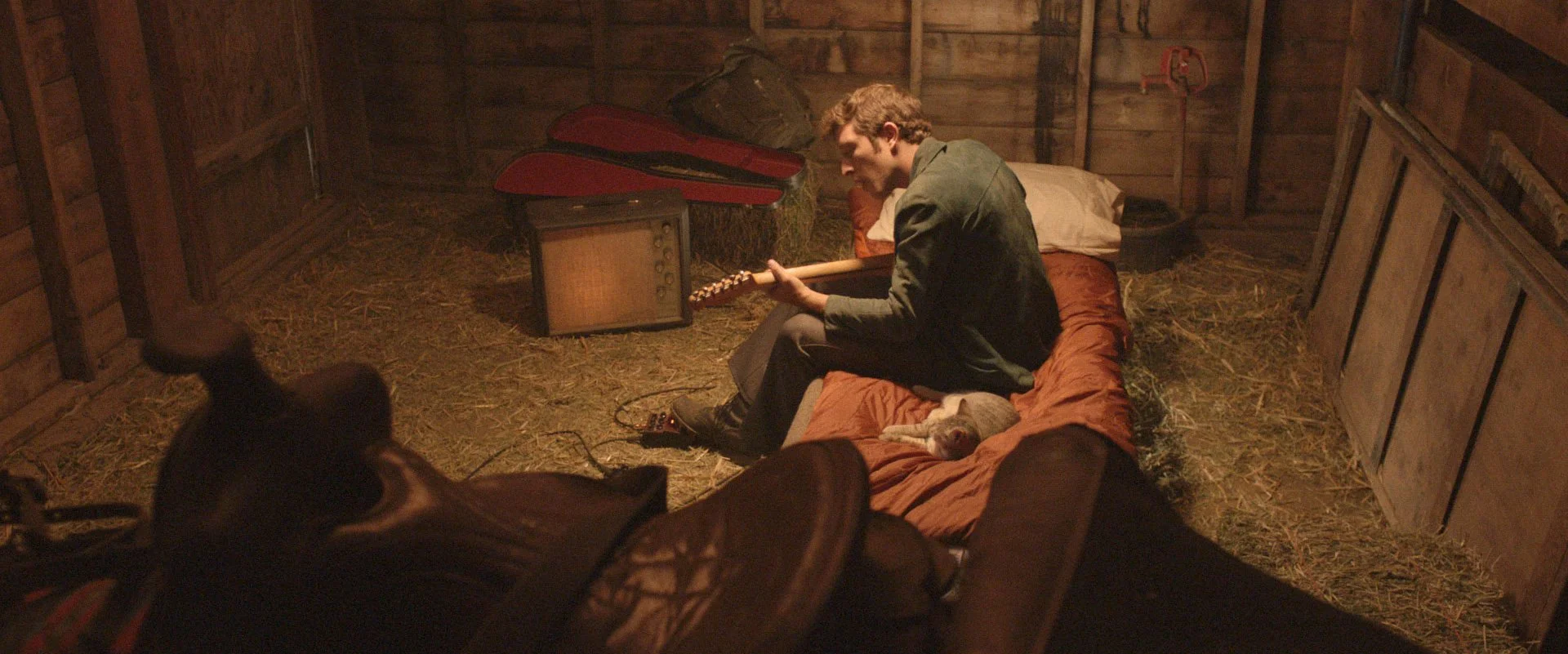 Gabriel Sunday sitting on a bed playing an electric guitar in a rustic barn with a cat lying next to him. There is an old radio, a guitar case, Analogman guitar pedal and hay on the ground. Directed by Jason Corgan Brown.
