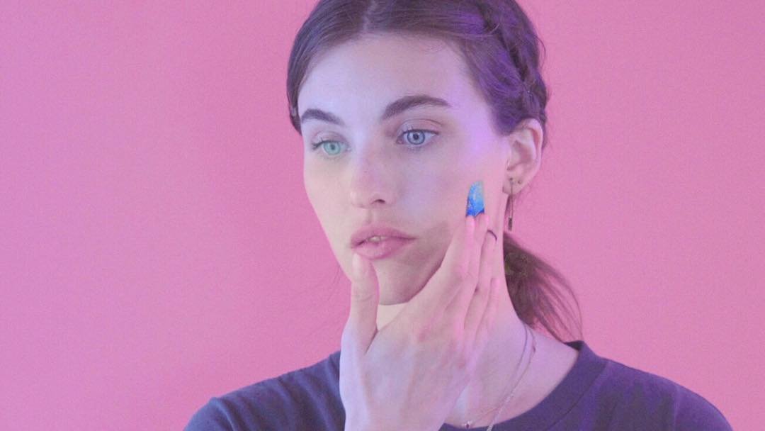 Young woman touching her face with her fingers against a pink background, with purple lighting on her hair.