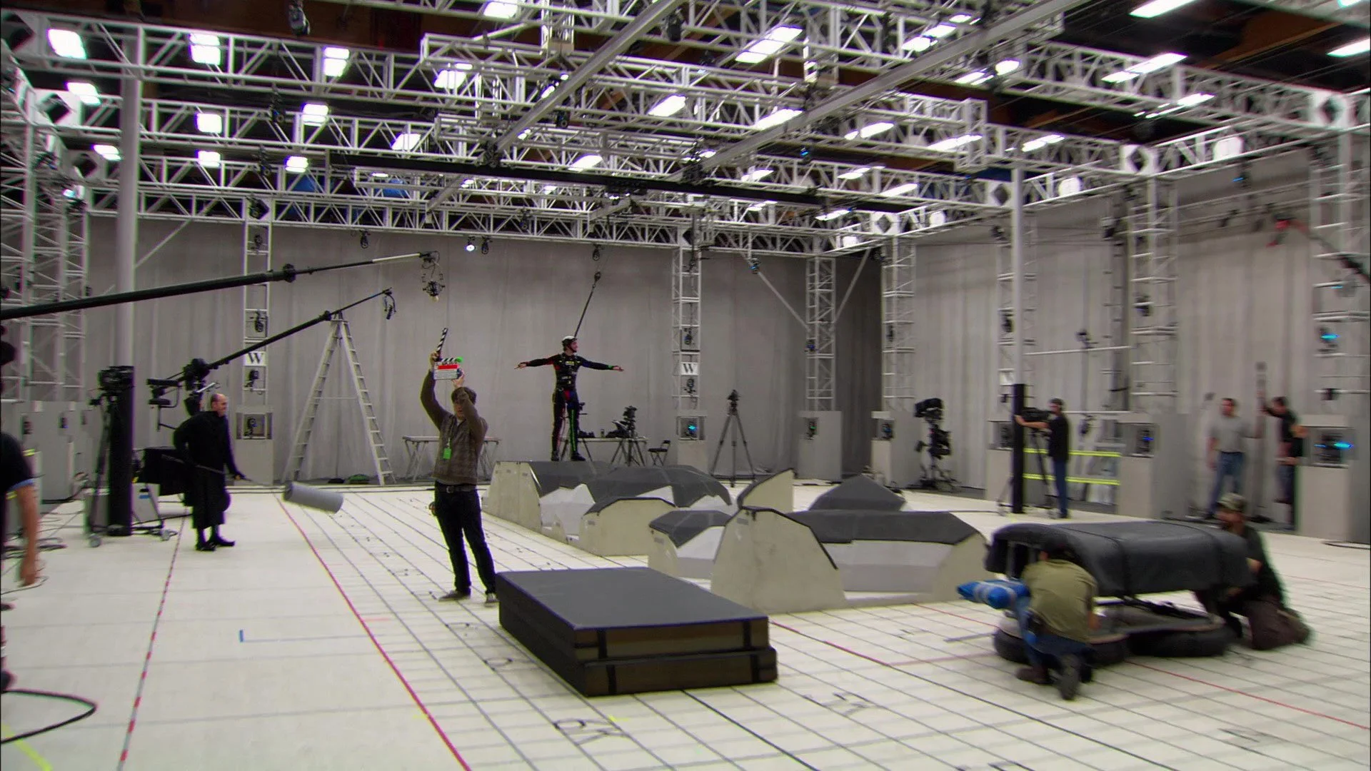 Behind the scenes of a motion capture studio with a person on a motion capture suit standing on a platform, crew members and cameras set up around the room, with a grid marked on the floor for positioning.
