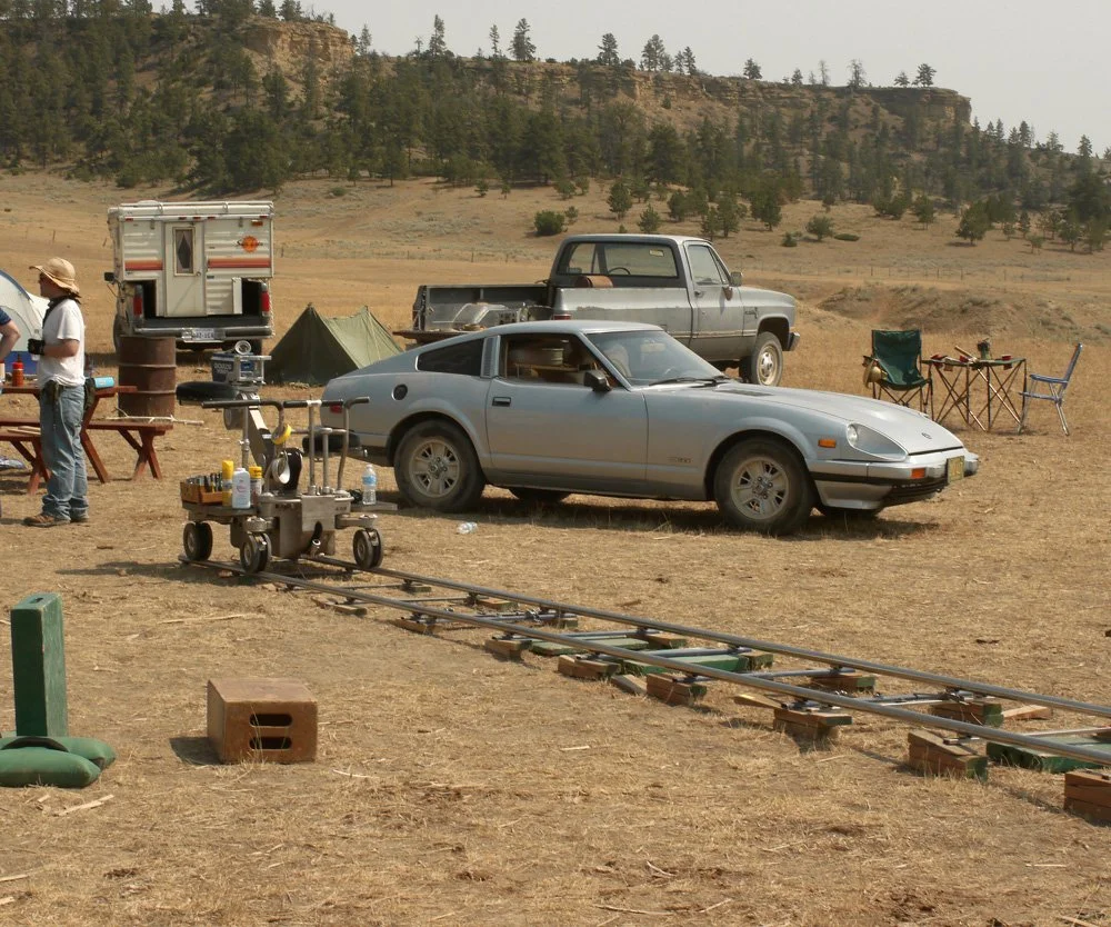 A dry, open outdoor setting with vintage and modern cars, camping gear, folding chairs, and people preparing for a film or photo shoot.