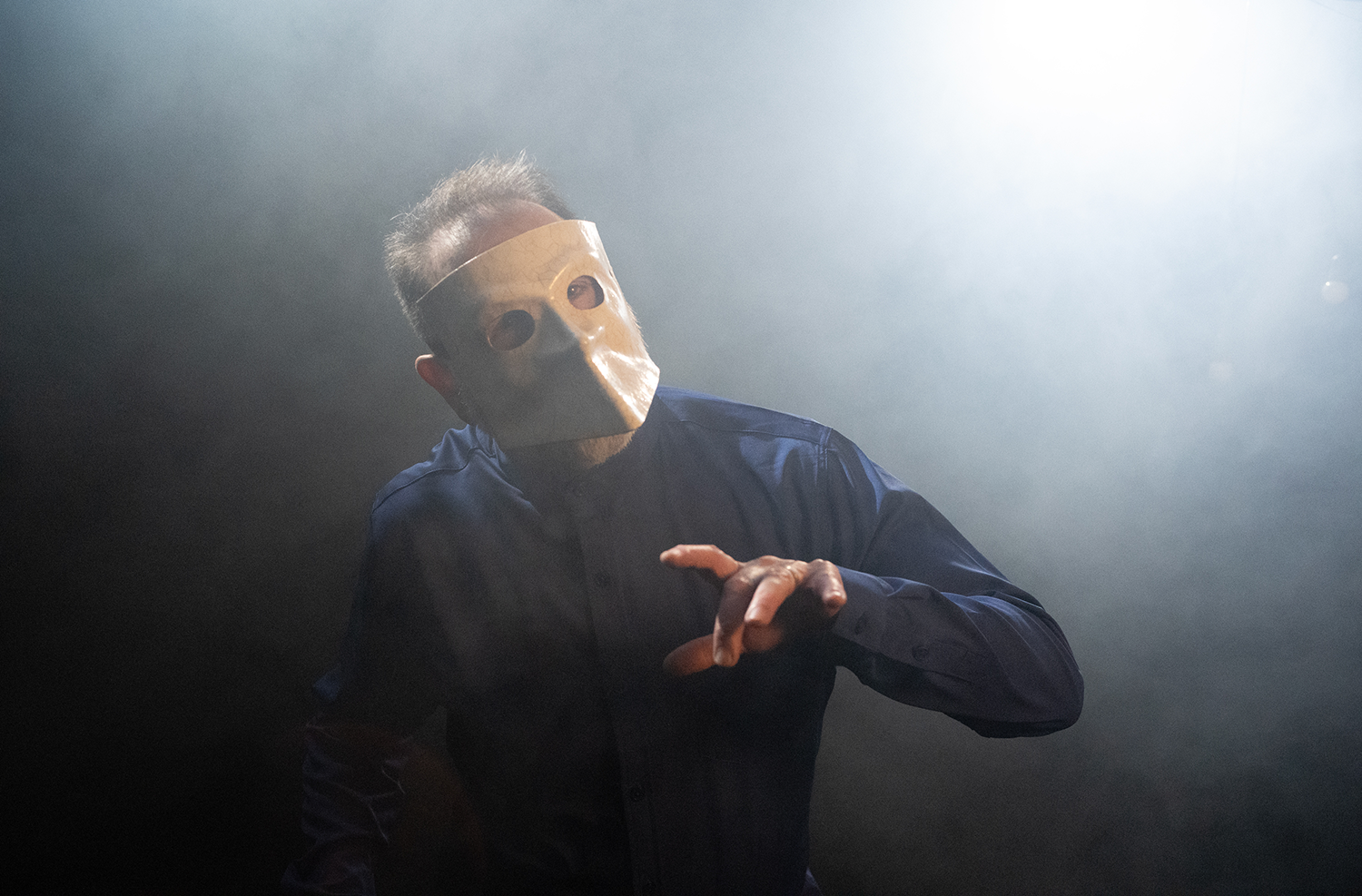 Joel Gori wearing a paper mask that resembles a human face, dressed in a dark blue shirt, is reaching out with one hand in a dimly lit environment with fog or smoke. Directed by Jason Corgan Brown. 