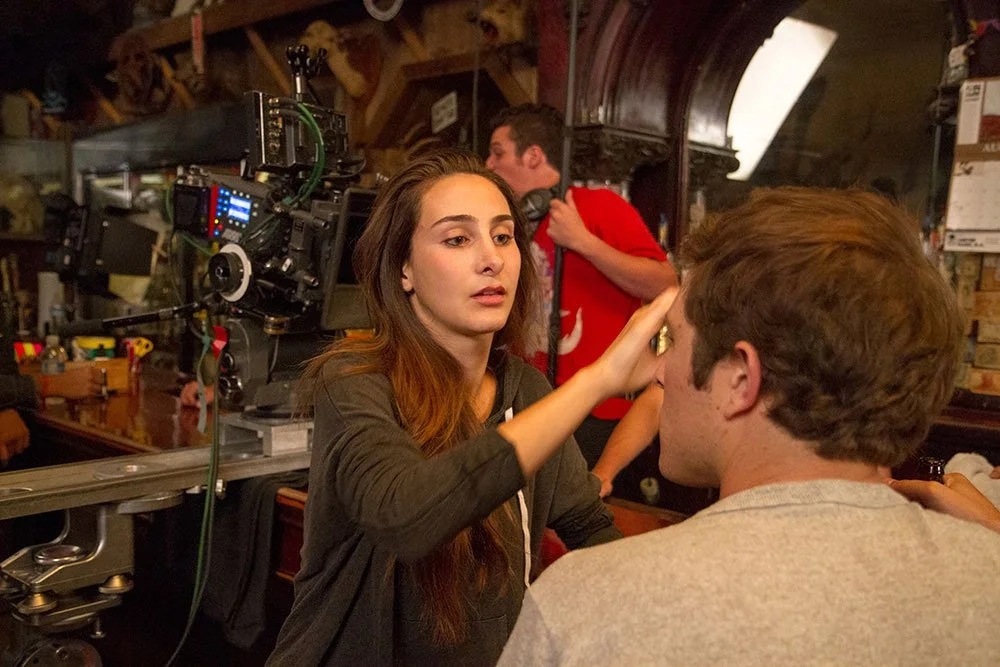 Sasha Glasser applies makeup or touches up a person's face on a Corgan Studio film set, with a camera and crew in the background. Directed by Jason Corgan Brown. 