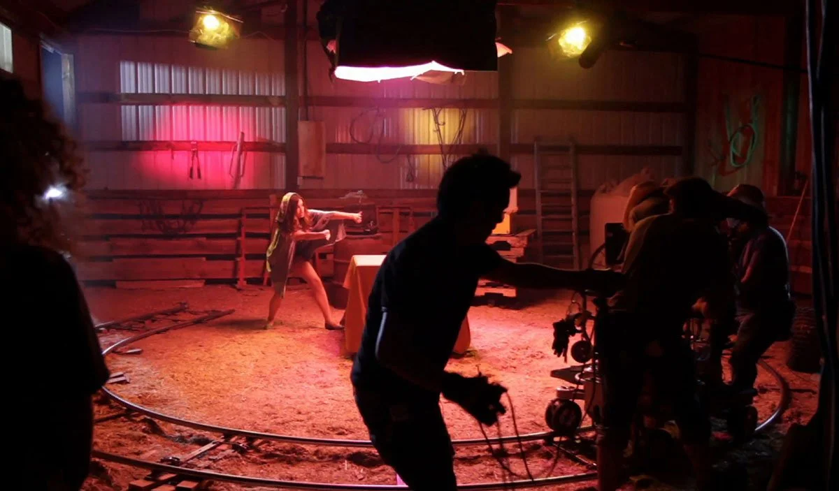 Behind-the-scenes photo on a film set shows a woman in costume performing a stunt, with crew members working around her, and the setup of lights and equipment in a barn-like structure.