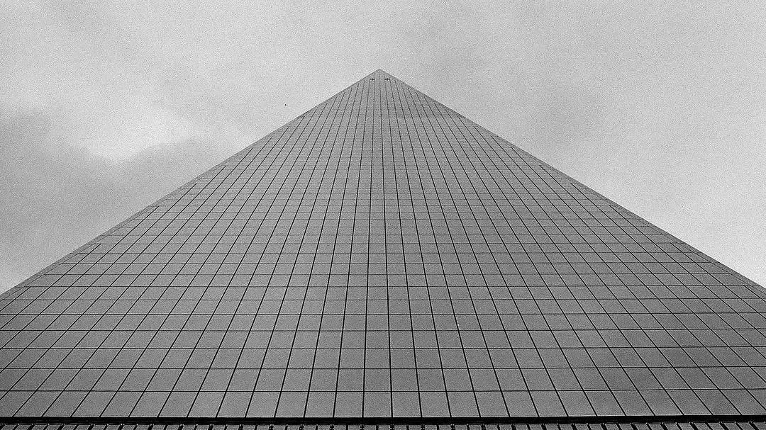 One World Trade Center, Freedom Tower in New York City.  A tall skyscraper with a pyramidal shape, covered in glass windows, seen from below against a cloudy sky. Film photography by Jason Corgan Brown. 