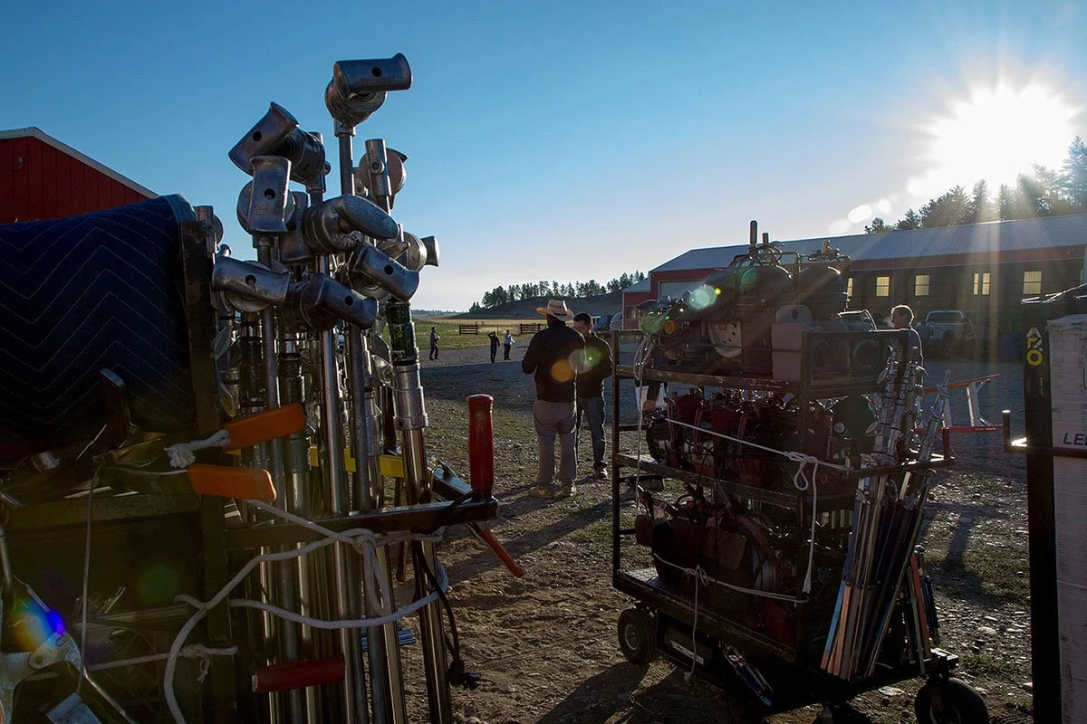 A collection of movie or camera equipment, including several lights, tripods, and camera gear, set up outdoors on a sunny day with a barn and people in the background.