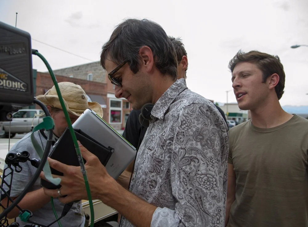 Group of people standing outdoors on a cloudy day, with Jason Corgan Brown looking at a tablet device, and others surrounding him, near equipment and a vehicle in the background.
