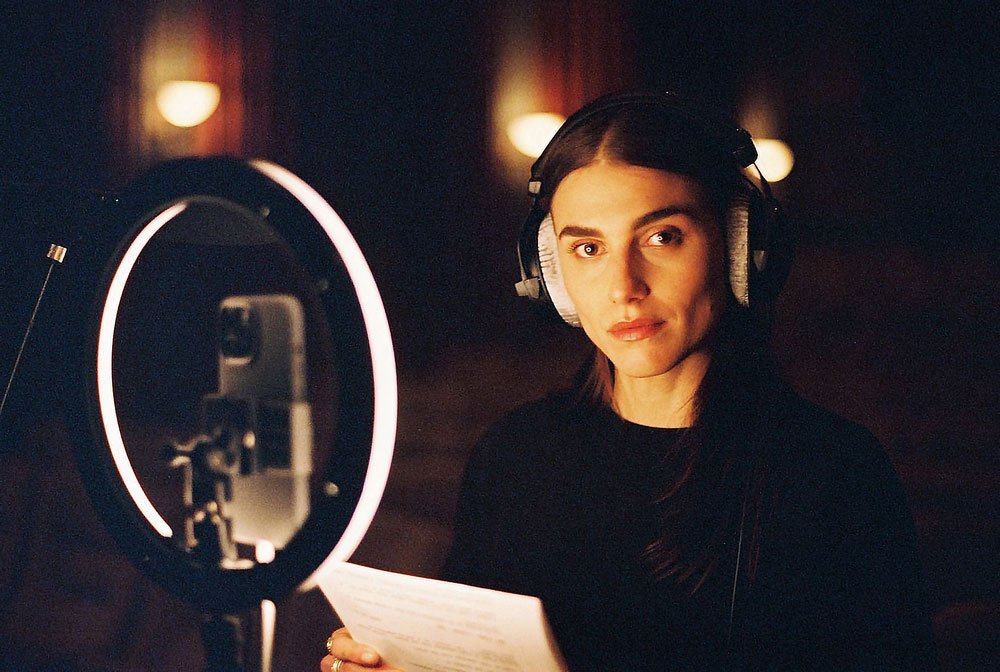 Langley Fox Hemingway wears headphones, standing on a film set with Jason Corgan Brown for Corgan Studio with a microphone and a pop filter, holding a script.