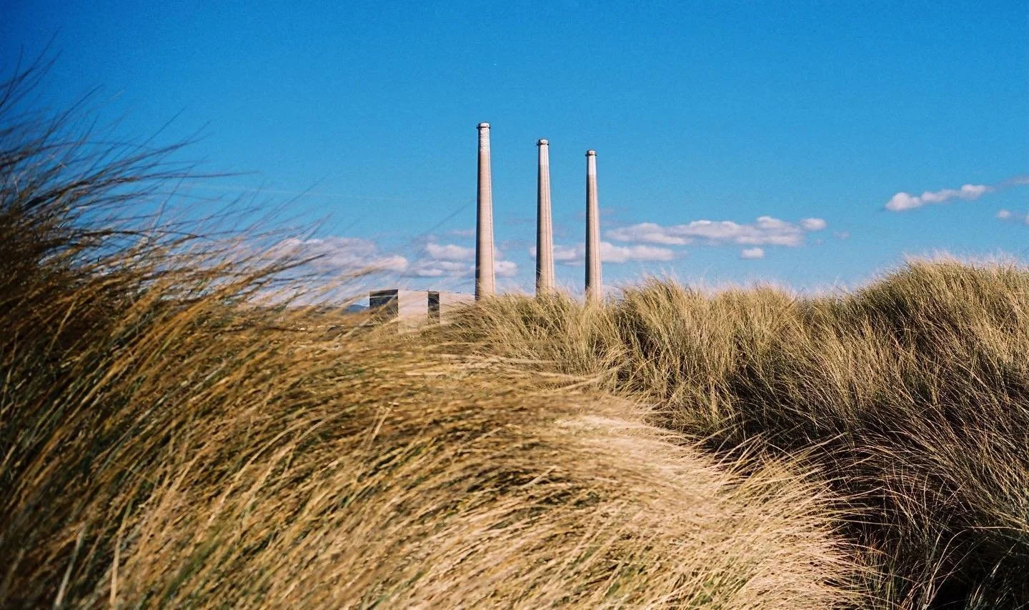 Tall grass on a sandy path with an industrial building and three smokestacks in the background under a partly cloudy blue sky. Film photography by Jason Corgan Brown. 