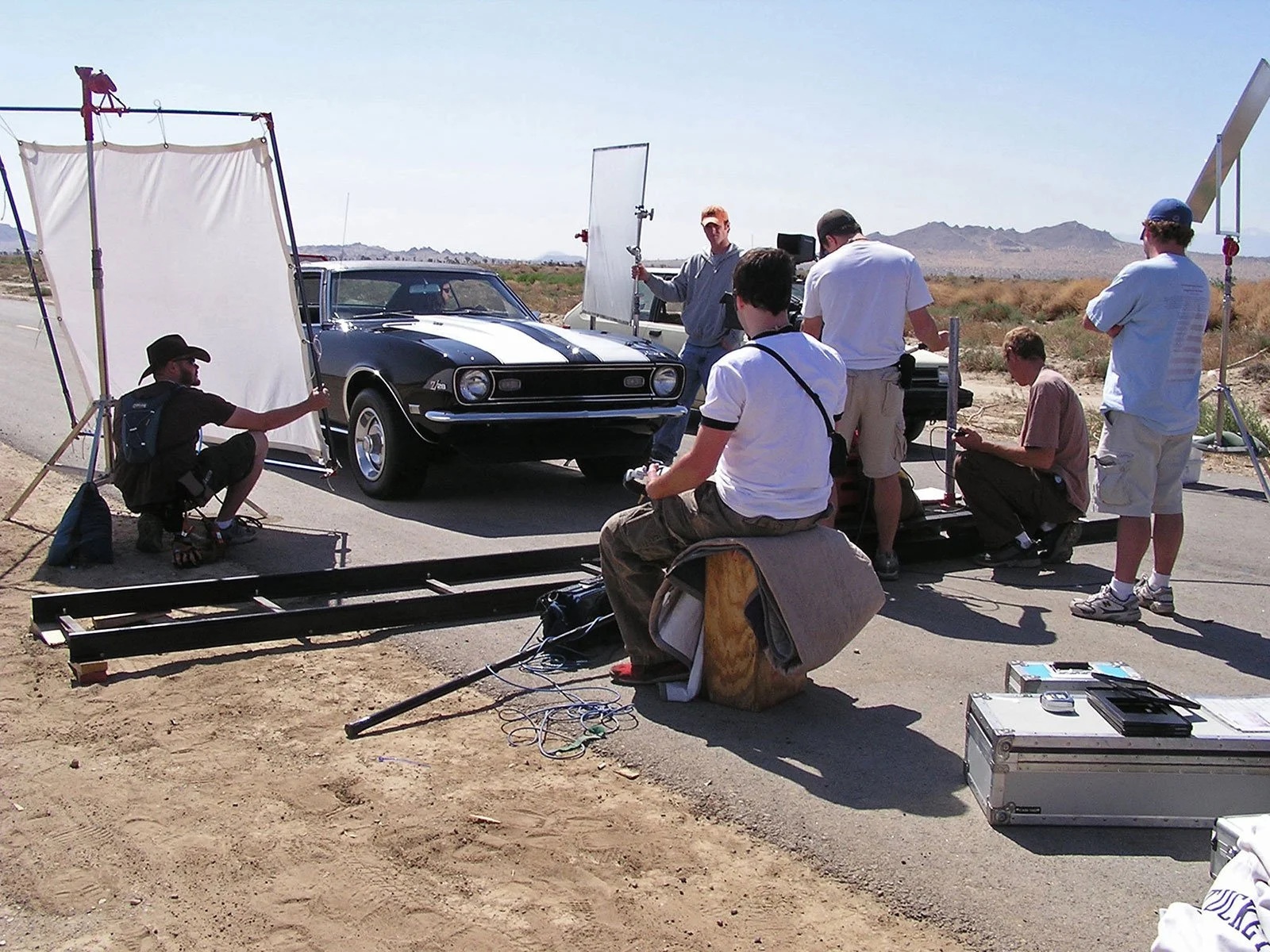 Behind-the-scenes view of a film crew filming a black vintage Ford Mustang with a white racing stripe in a desert setting, with equipment and crew members preparing for the shot. Directed by Ryan Nelson, Produced by Jason Corgan Brown. 