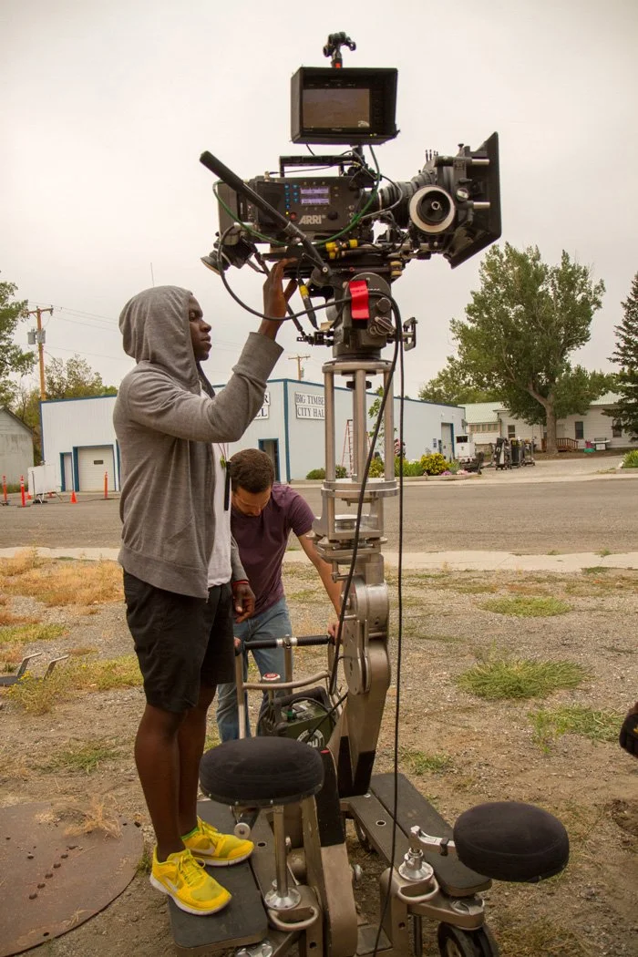 Two men operating a large professional video camera outdoors, with one standing and adjusting the camera and the other behind it, in front of a small urban commercial area with trees and buildings.
