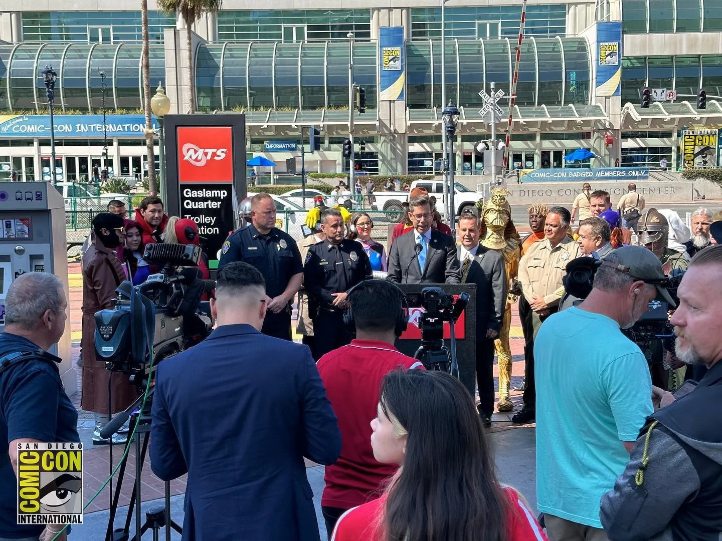 A group of people, including police officers and individuals in costume, are gathered outside the San Diego Convention Center during Comic-Con International. There are cameras and microphones set up, indicating a press conference or public event.