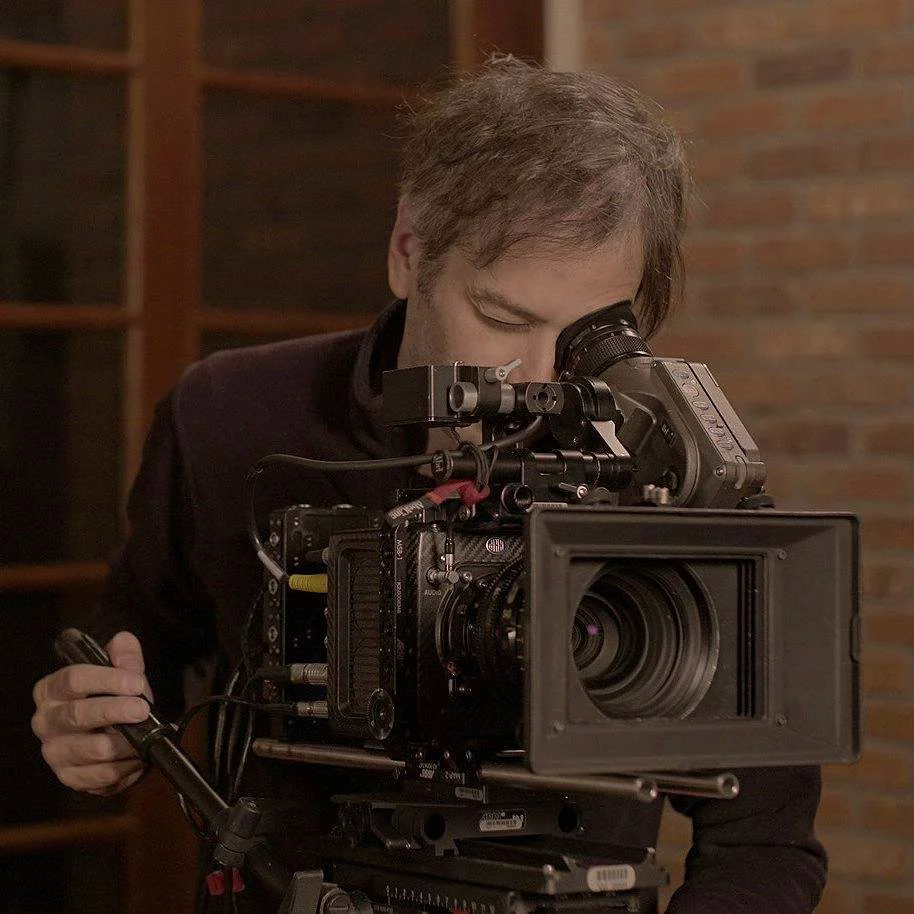A man operating a professional film or video camera indoors, with a brick wall in the background.