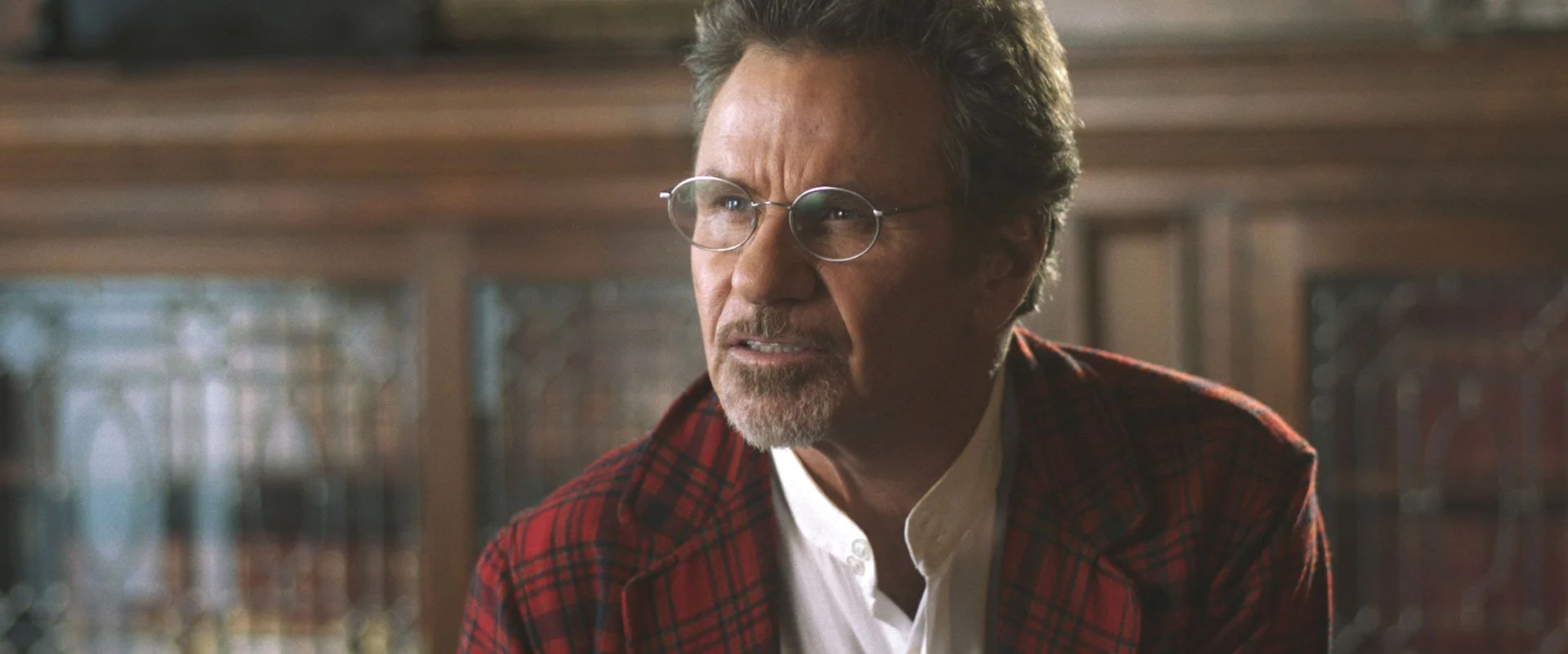 A middle-aged man with glasses and gray hair, wearing a red and black plaid jacket over a white shirt, appears to be indoors in a setting with wooden furniture and decorative glass windows.