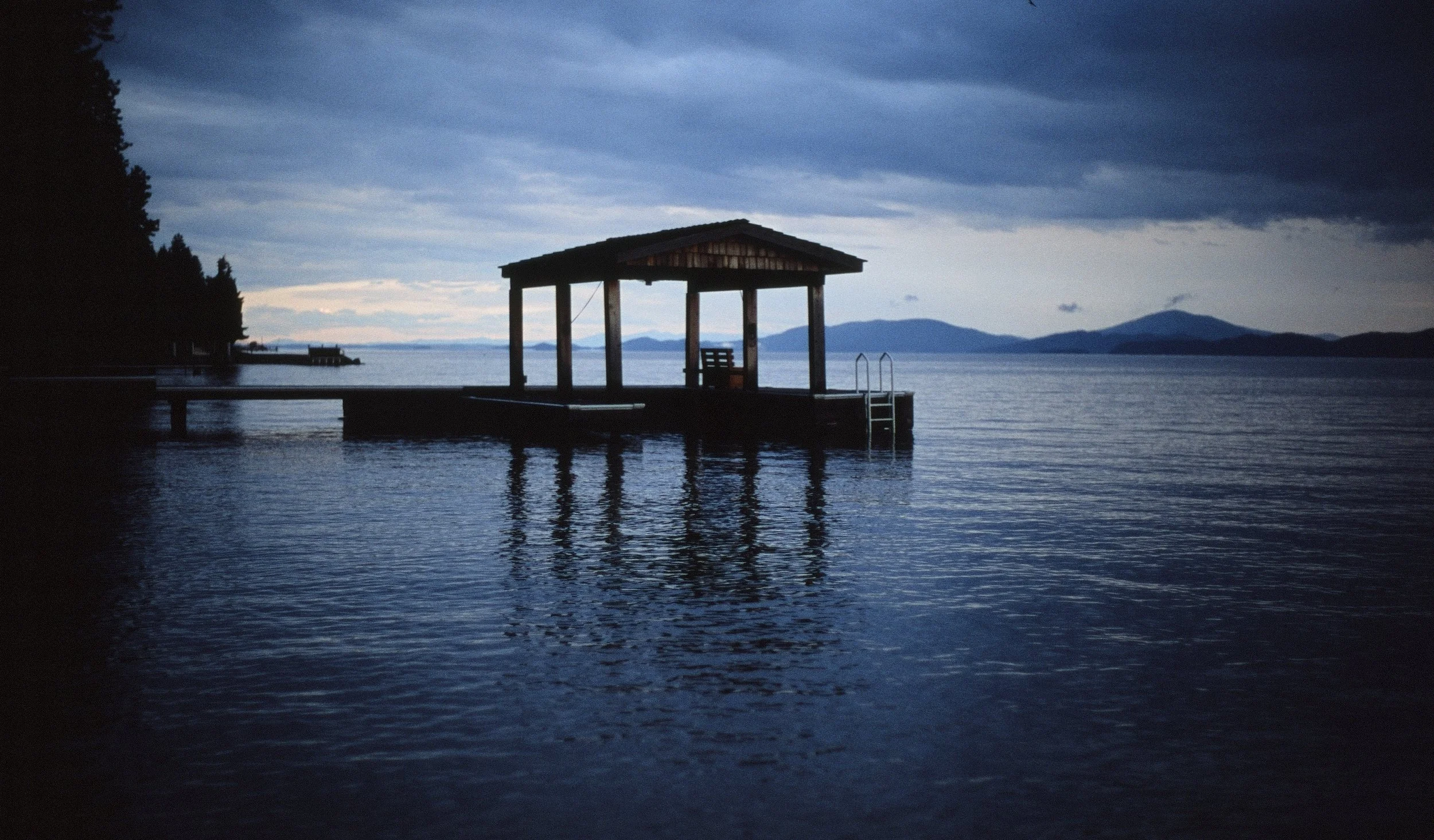 A wooden pier extends into a calm lake with a small covered shelter at the end. The scene is viewed during dusk or early evening, with dark clouds overhead and distant mountains on the horizon.