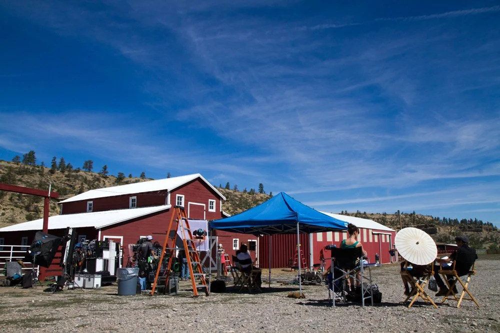 Filming crew setting up scene in front of a red barn on a sunny day, with people seated under a blue canopy and a white umbrella.