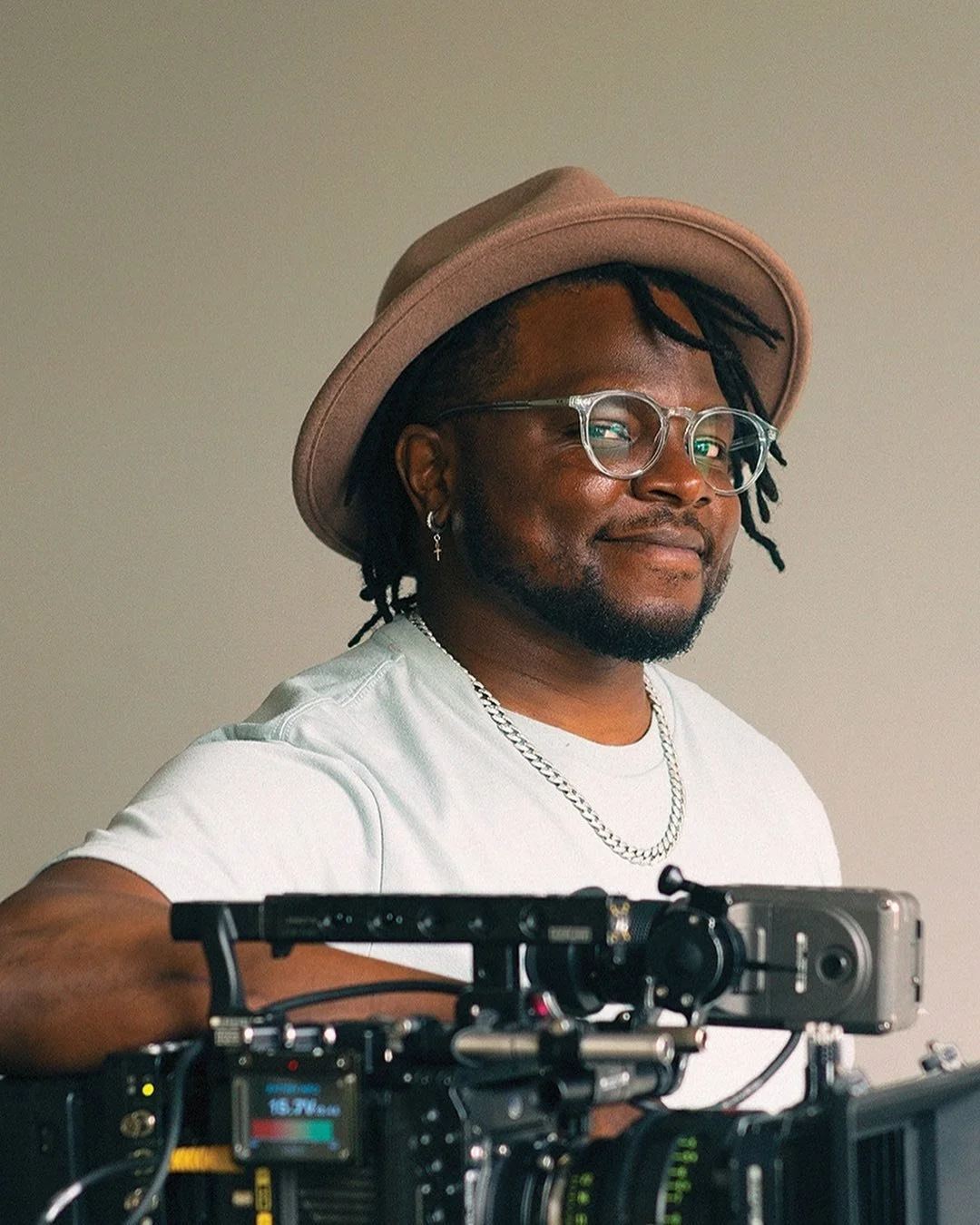 Bruce Francis Cole wearing a tan hat, clear glasses, silver earrings, and a silver chain, taking a selfie with a camera.