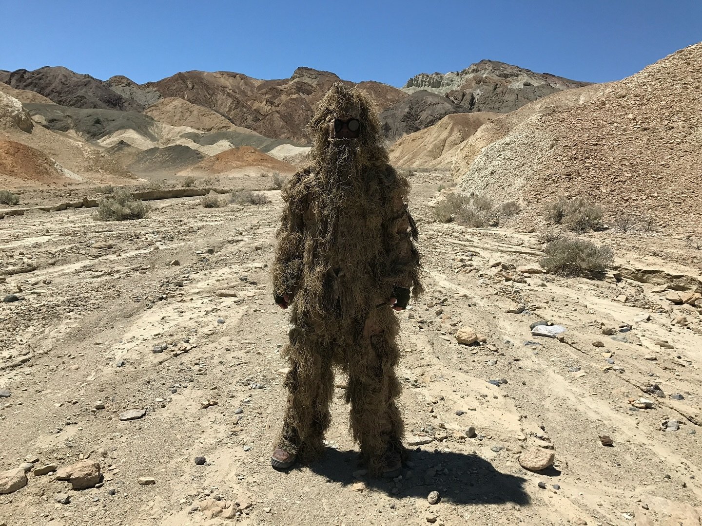 Person dressed in a full ghillie suit standing in a desert landscape film set for Corgan Studio with mountains in the background.