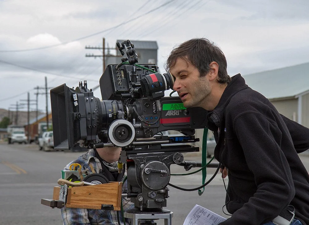 Jason Corgan Brown looking into a professional film camera on a street, with houses and parked cars in the background.