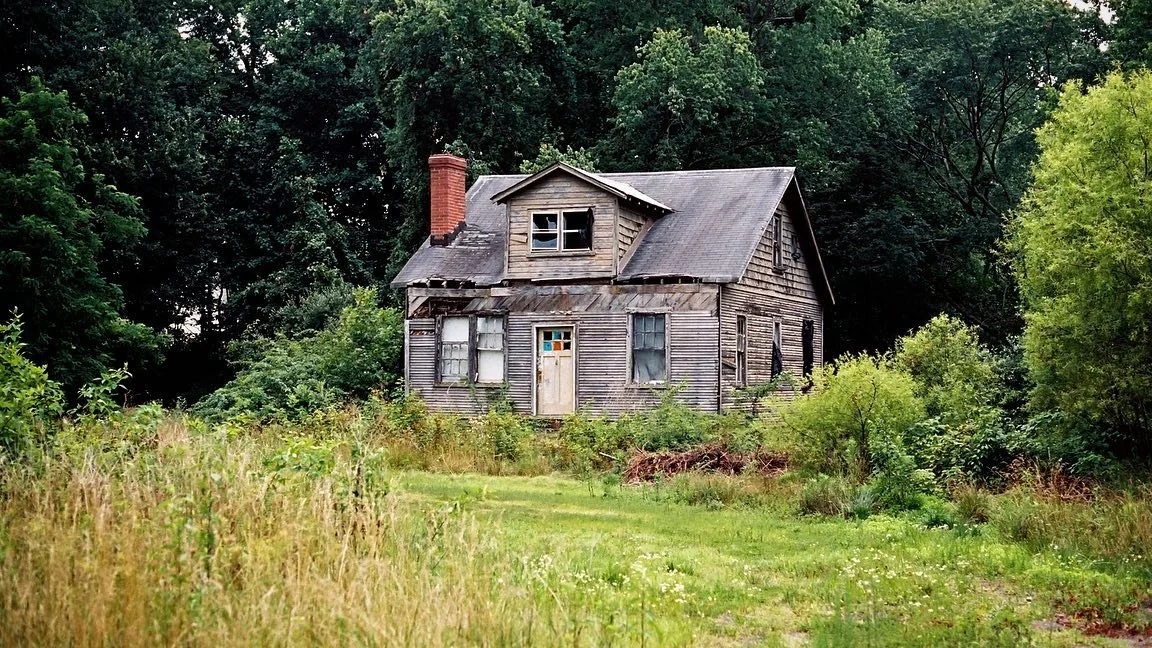 An old, abandoned house with peeling siding and broken windows, surrounded by overgrown grass and trees in a rural setting.