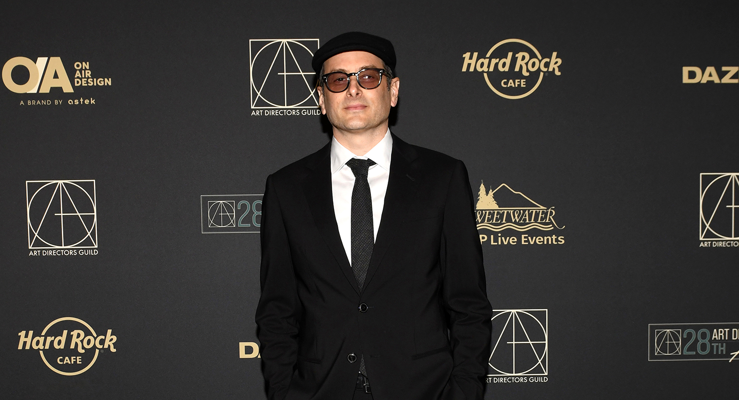 Man in a black suit, white shirt, black tie, and black hat standing against a black backdrop with logos including Hard Rock Cafe and Art Directors Guild.
