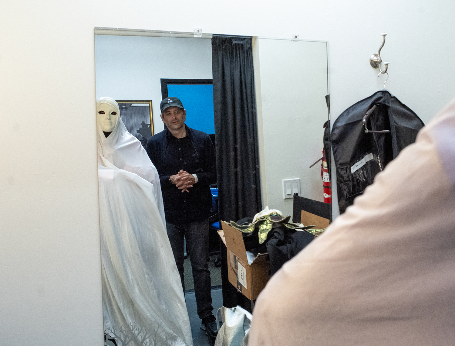 A person in white costume with a mask stands next to a man in a black cap, both reflected in a mirror, with a box of fabric or costumes and a large black bag nearby. Directed by Jason Corgan Brown. 